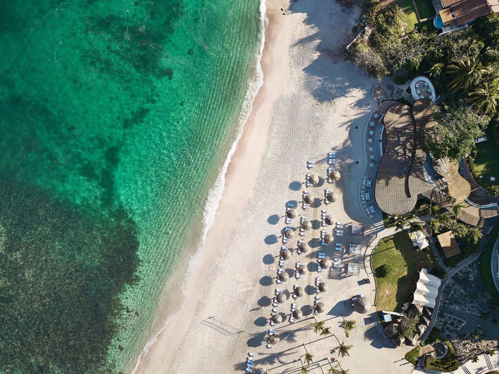 An aerial view of a beach resort showing turquoise ocean water, sandy beach with lounge chairs and umbrellas, and a resort building with palm trees and greenery.