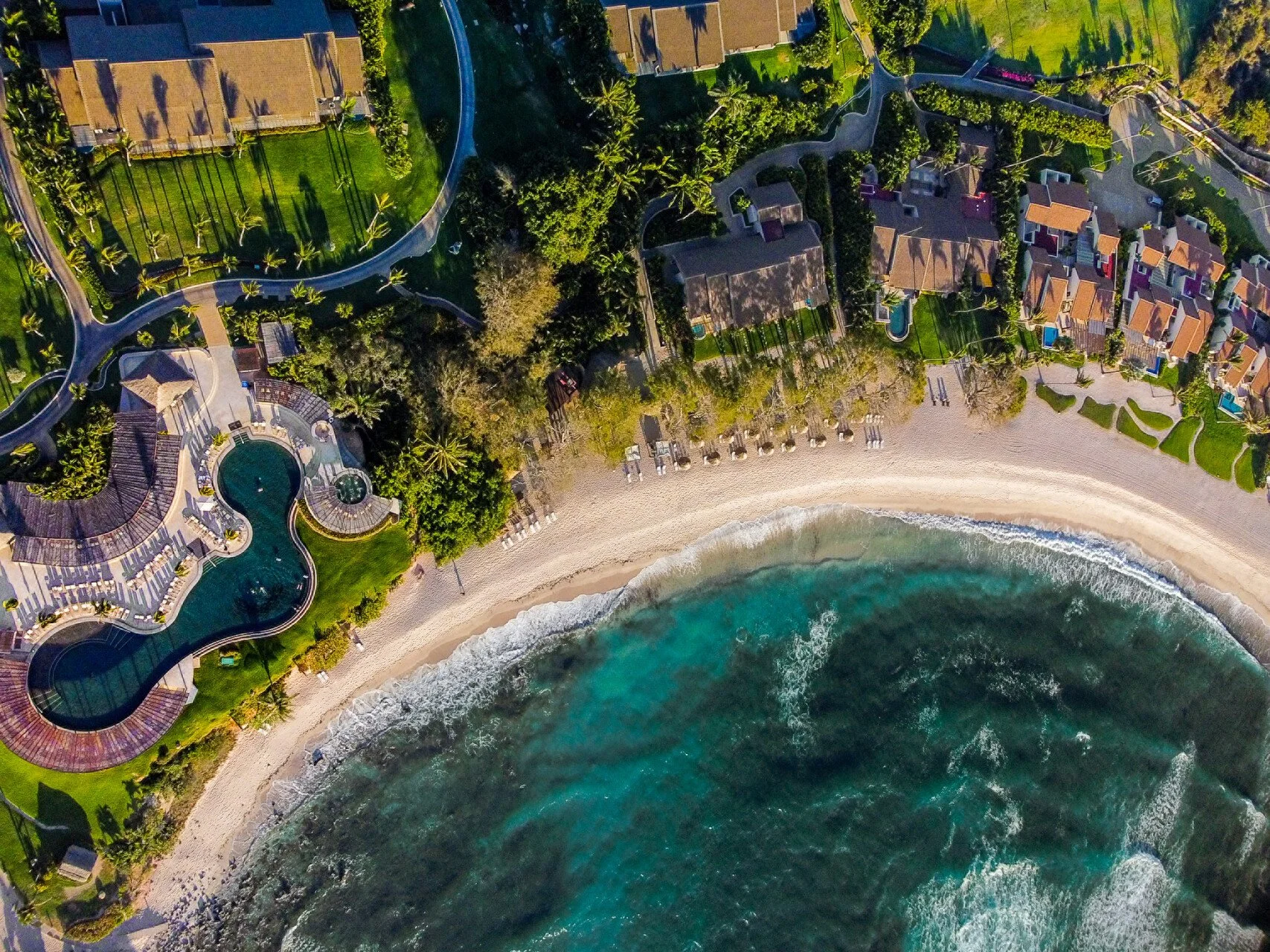 An aerial view of a beachfront resort with swimming pools, tropical greenery, and beachfront volleyball courts, adjacent to the ocean with waves and a sandy beach.