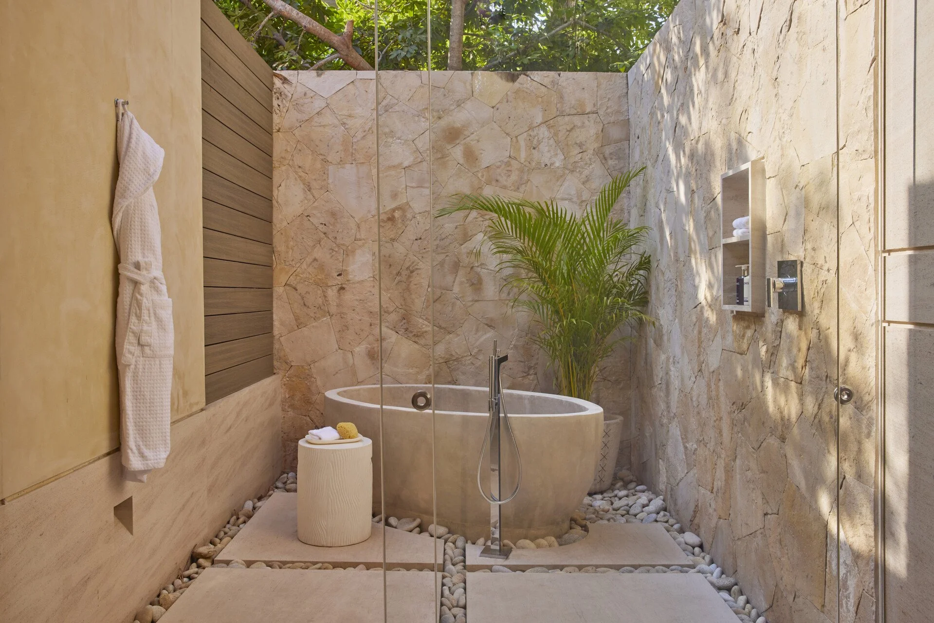Outdoor bathroom with beige stone walls, glass shower door, freestanding oval bathtub, a potted plant in the corner, and a small side table with towels and bath accessories.