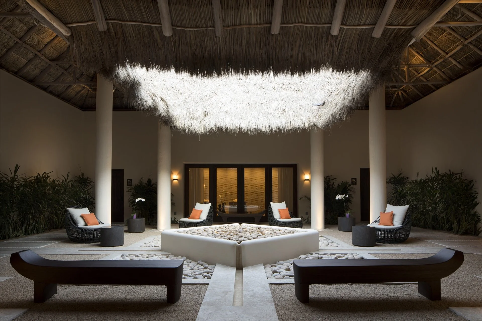 Indoor lounge area with wicker chairs, orange pillows, potted orchids, and a central white rock-filled square, under a thatched ceiling with wooden beams and ambient lighting.