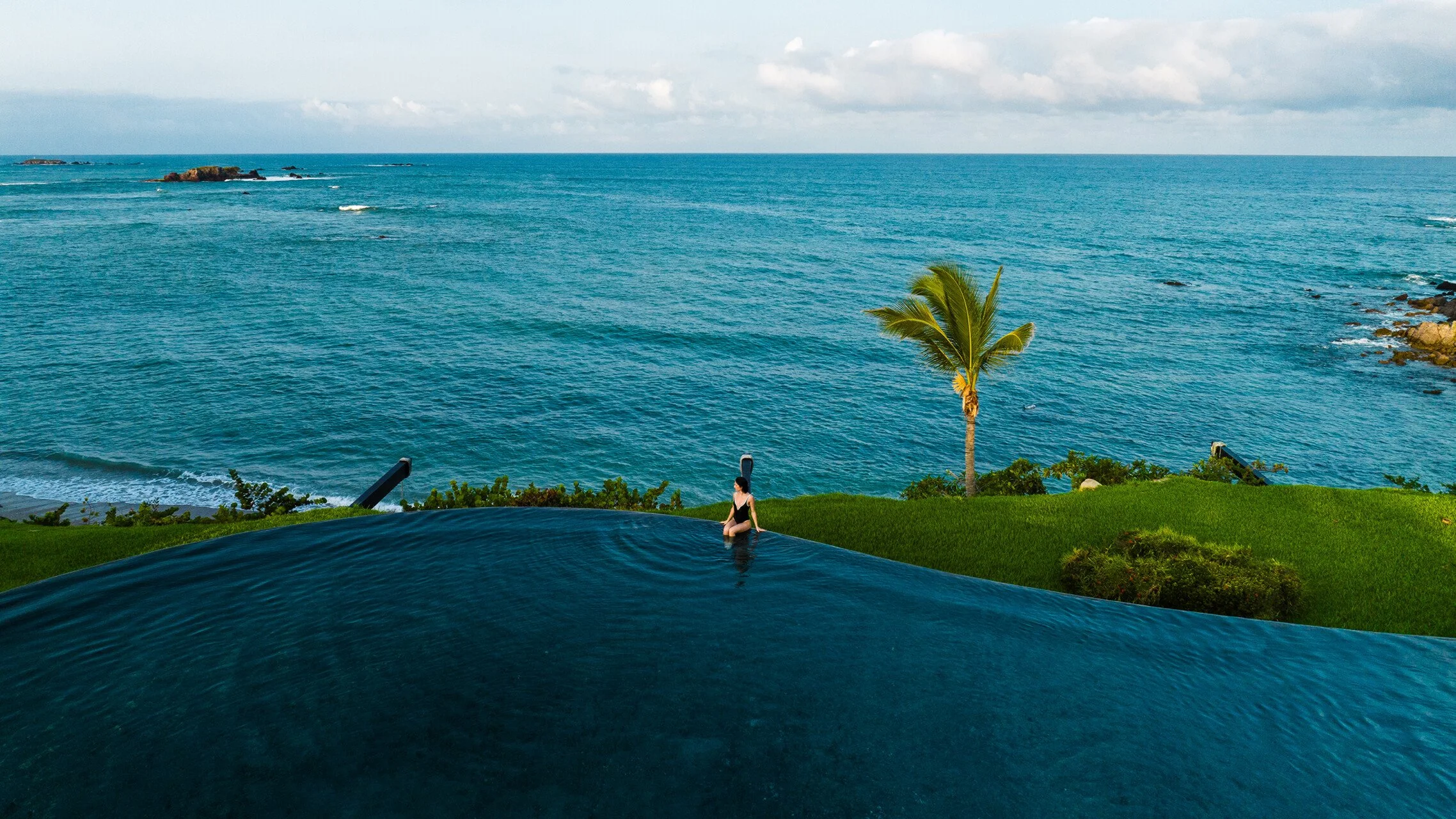 Person relaxing in an infinity pool overlooking the ocean with a palm tree and rocks in the background.