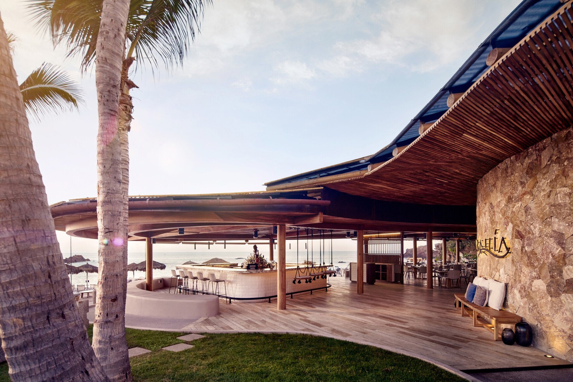 An outdoor bar and seating area at a beach resort, with palm trees, wooden decking, and the ocean in the background.