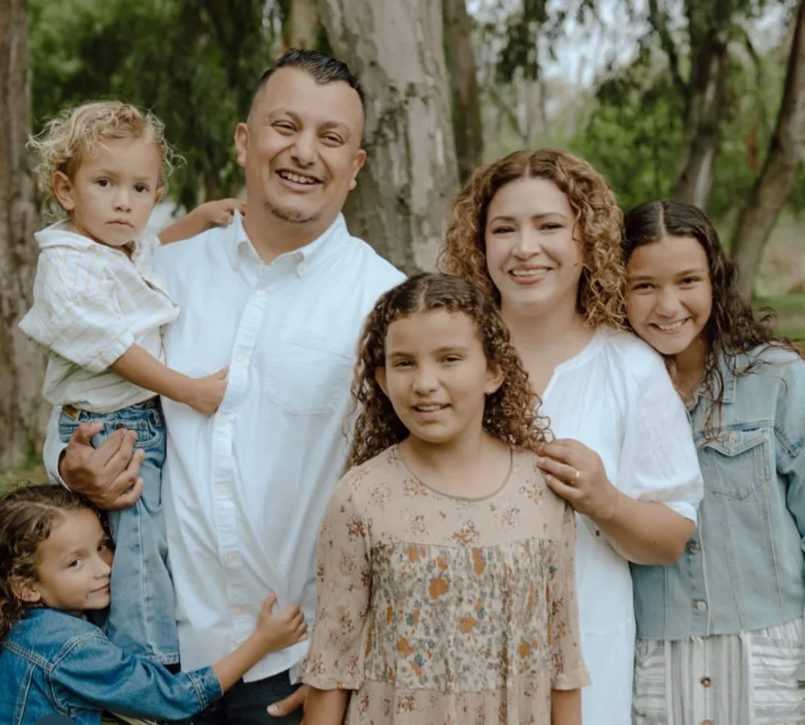Family of six standing outdoors near trees, smiling at camera. Two adults and four children, all dressed in casual clothing.