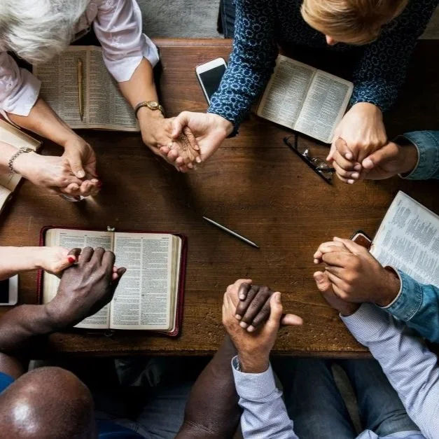 Group of diverse people holding hands in prayer around a wooden table with open Bibles and a smartphone.