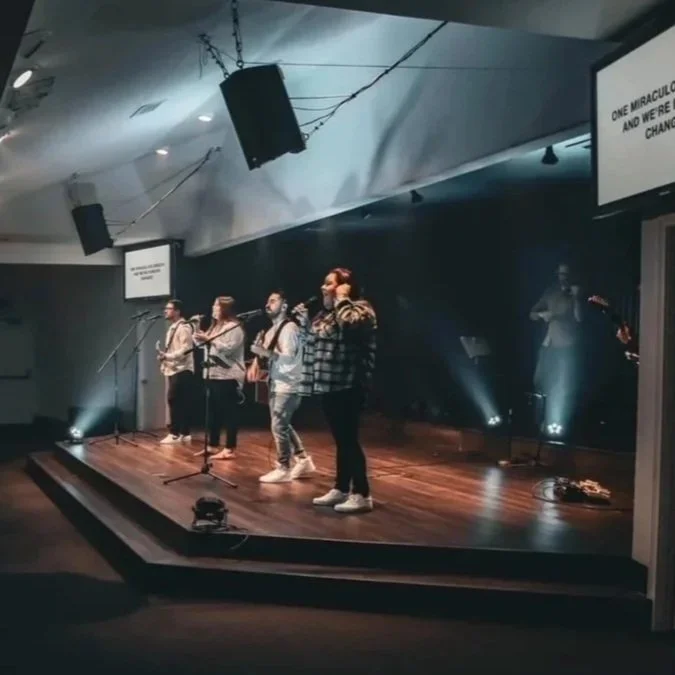 A band performing on stage with four members singing and playing instruments, in a dimly lit indoor venue with stage lights.