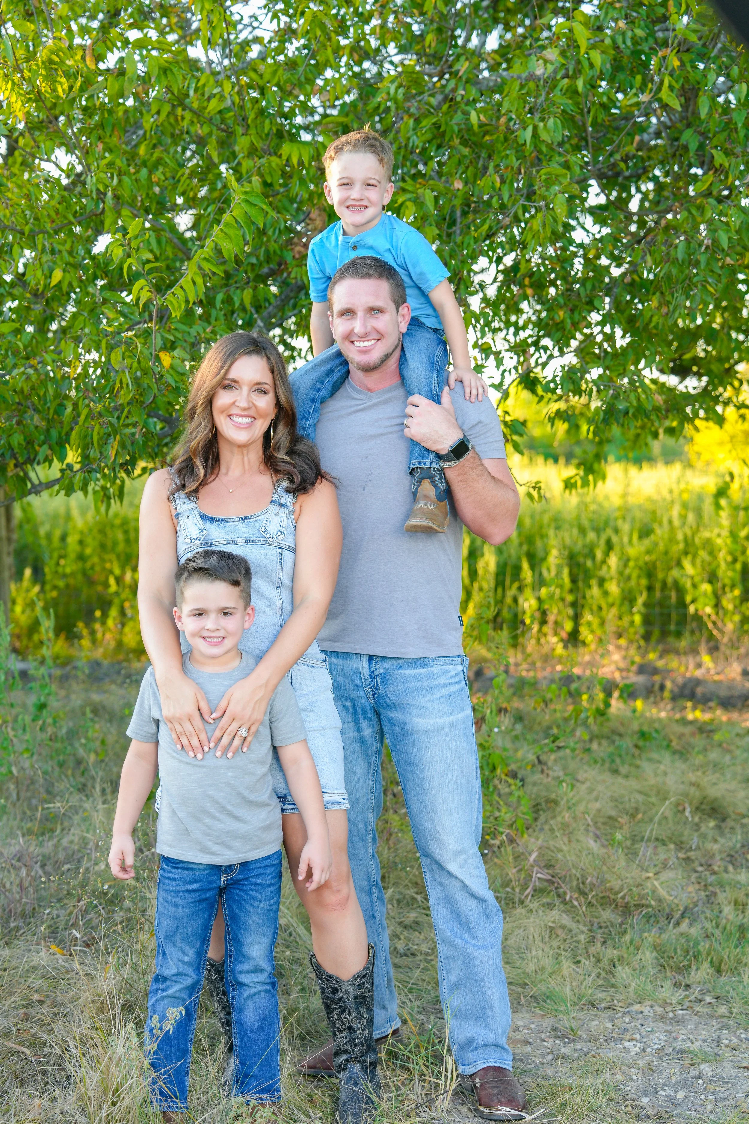 A family of four outdoors in a green, natural setting, with a tall man carrying a young boy on his shoulders, a woman standing beside him, and a young boy in front of her.