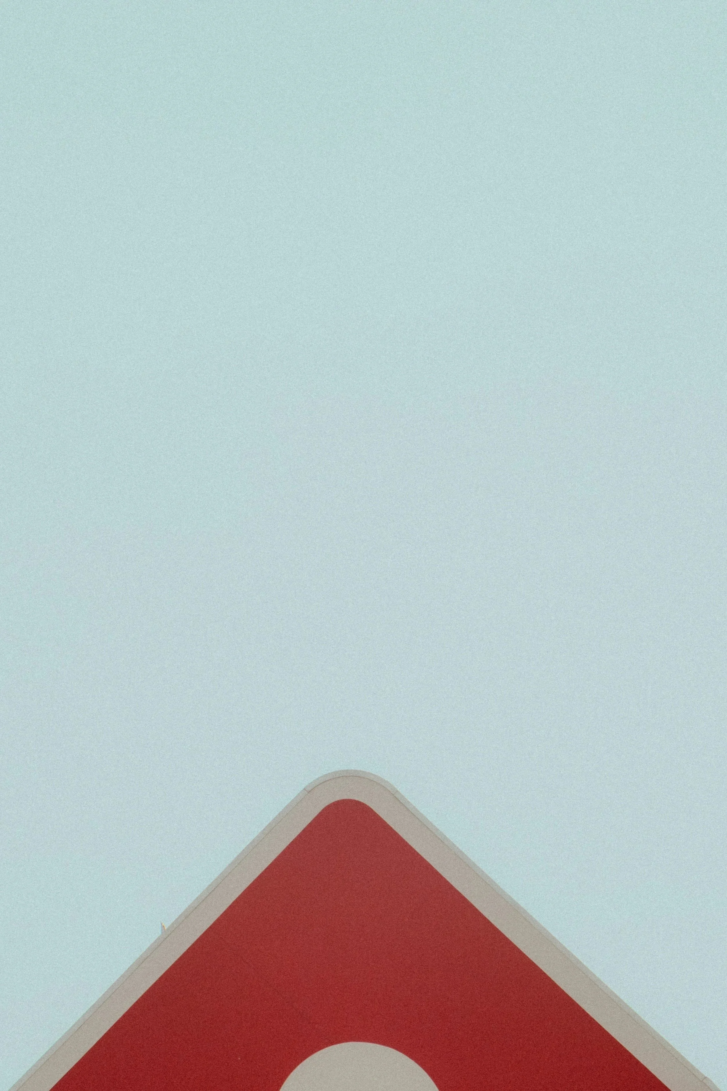 A red caution sign with a white circle at the bottom corner against a pale blue sky.