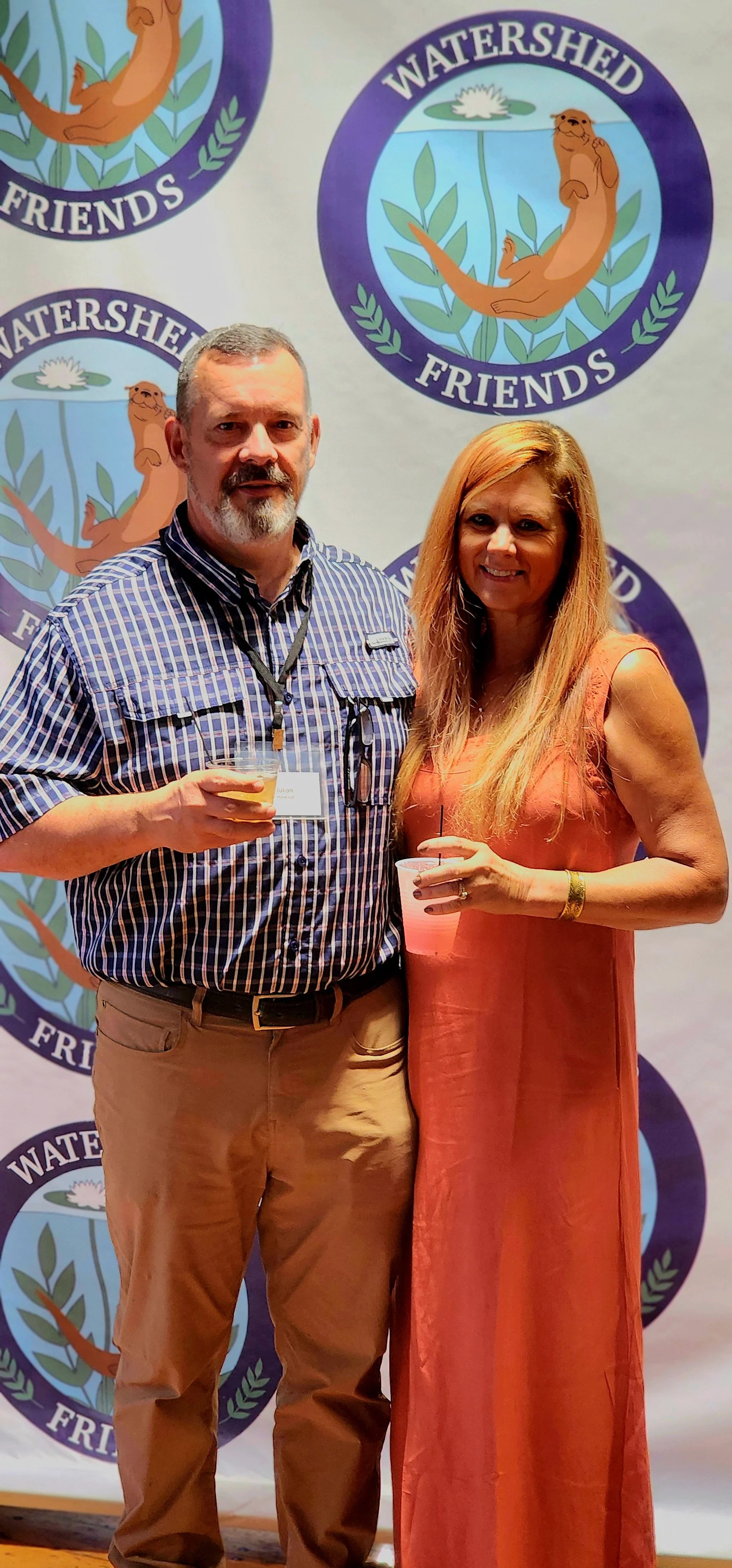 A man, wearing a blue and white plaid shirt and khakis, and a woman, wearing a coral dress, standing in front of a backdrop with the Watershed Friends logo