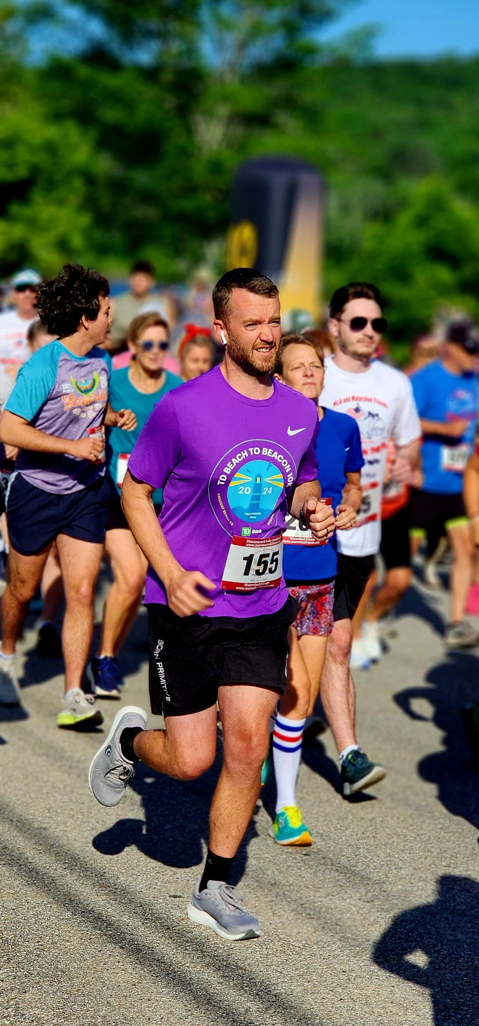 A man in a purple shirt and black shorts, running with a crowd of runners behind 