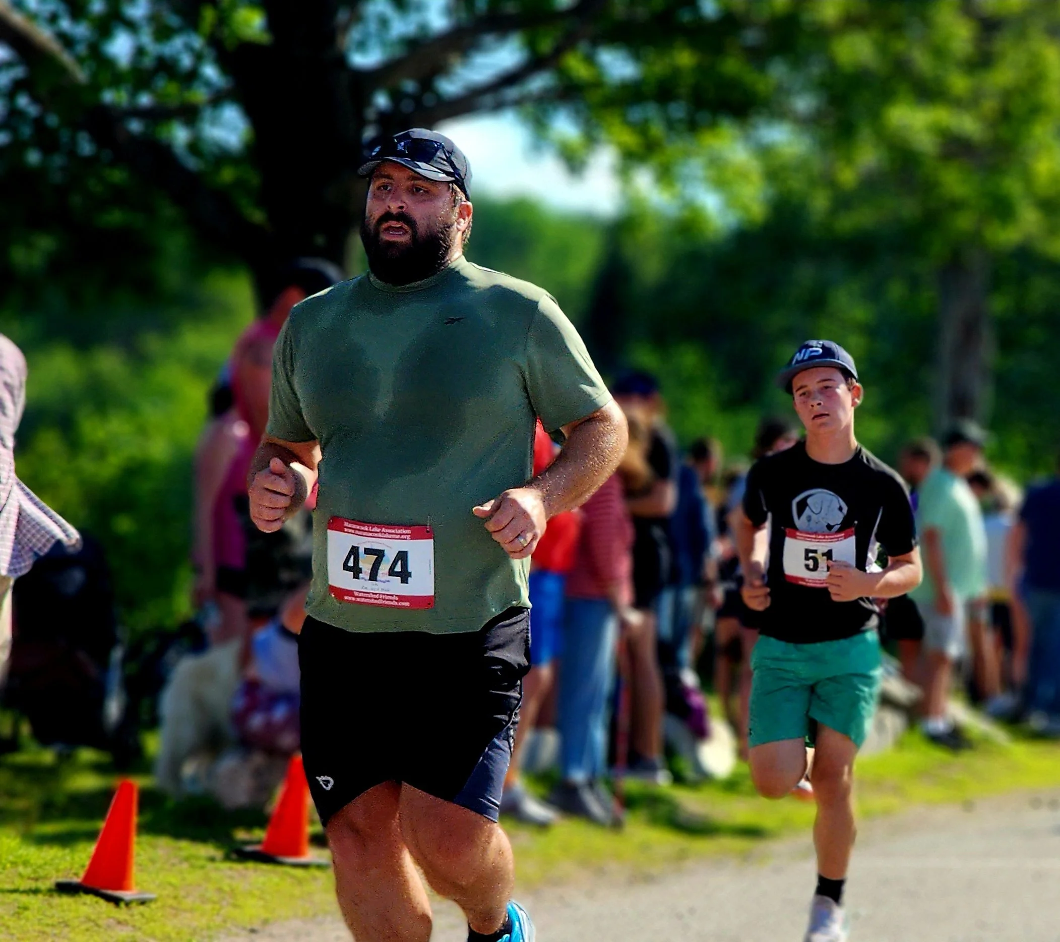 A man wearing a green t-shirt and black shorts running ahead of a boy wearing a black t-shirt and green shorts with a crowd standing in the background