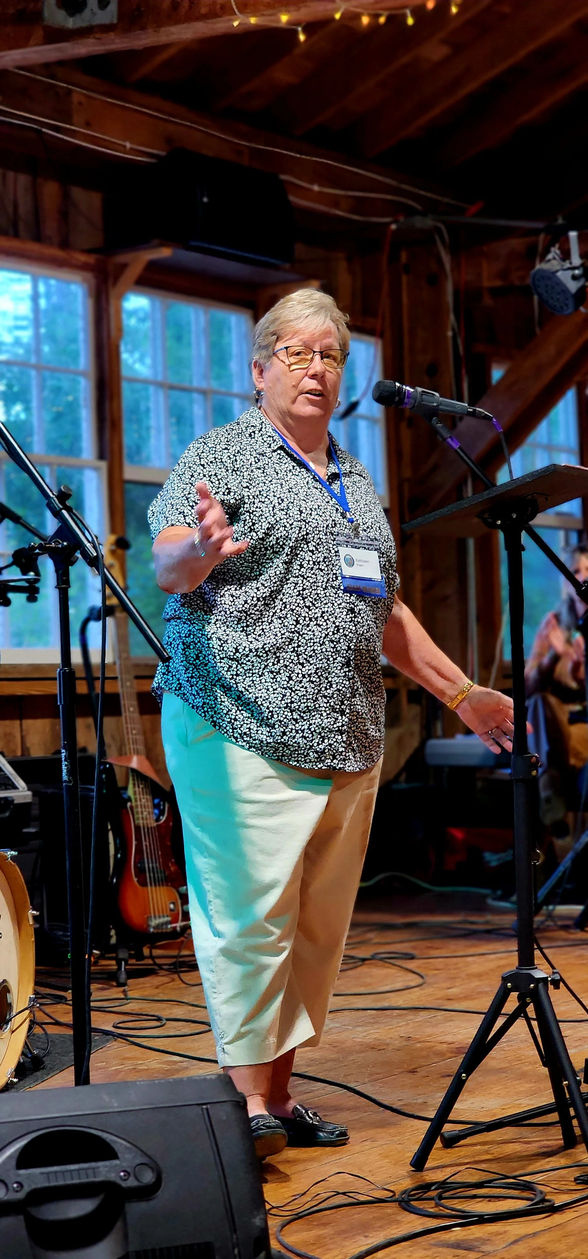 A woman, wearing a blue and white floral shirt and white pants, speaking into a microphone