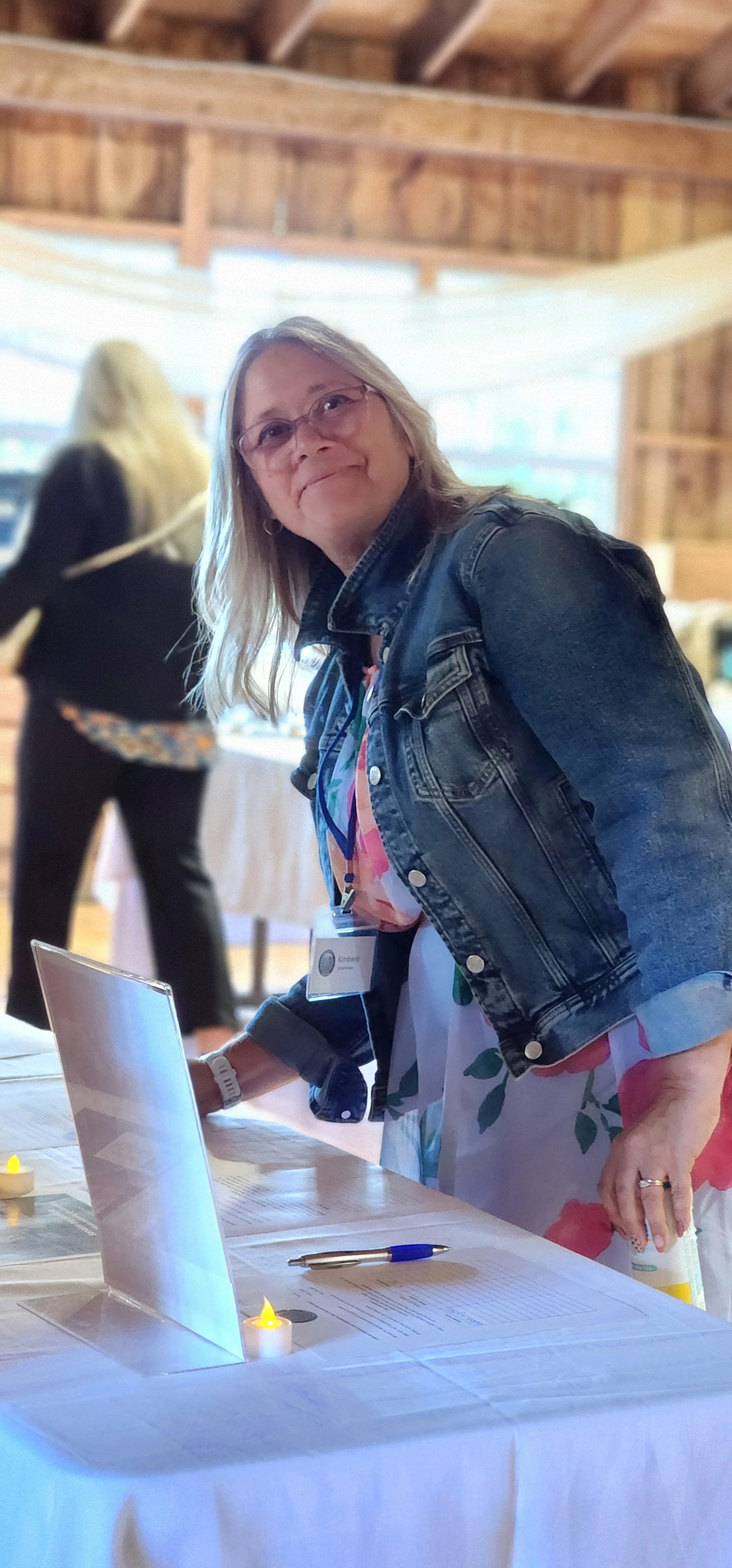 A woman, wearing a jean jacket over a floral patterned dress, leaning over a table and glancing at the camera