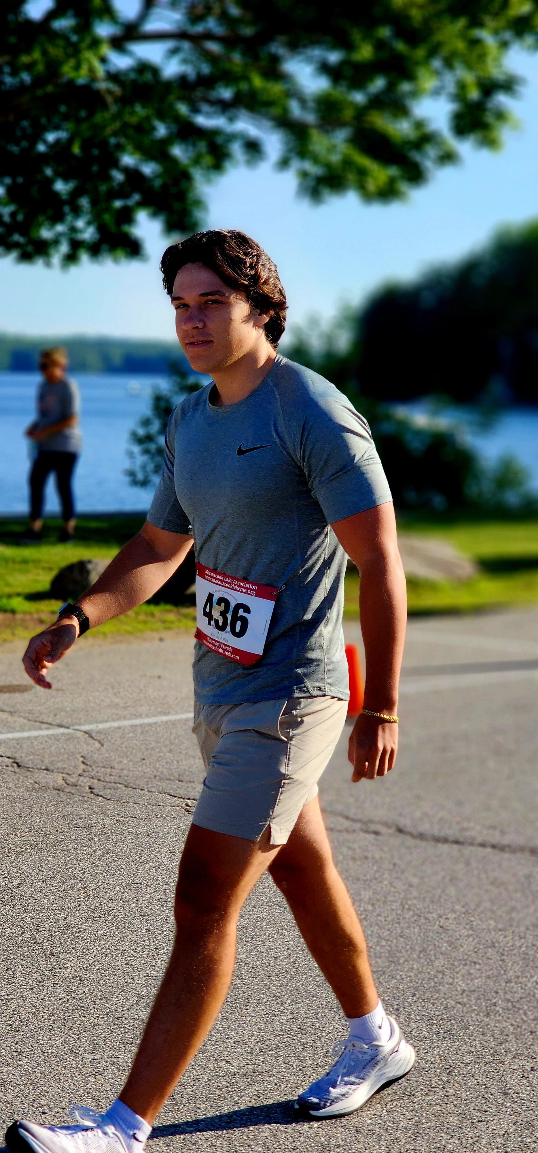 A man, wearing a grey t-shirt and grey shorts, walking with a lake in the background