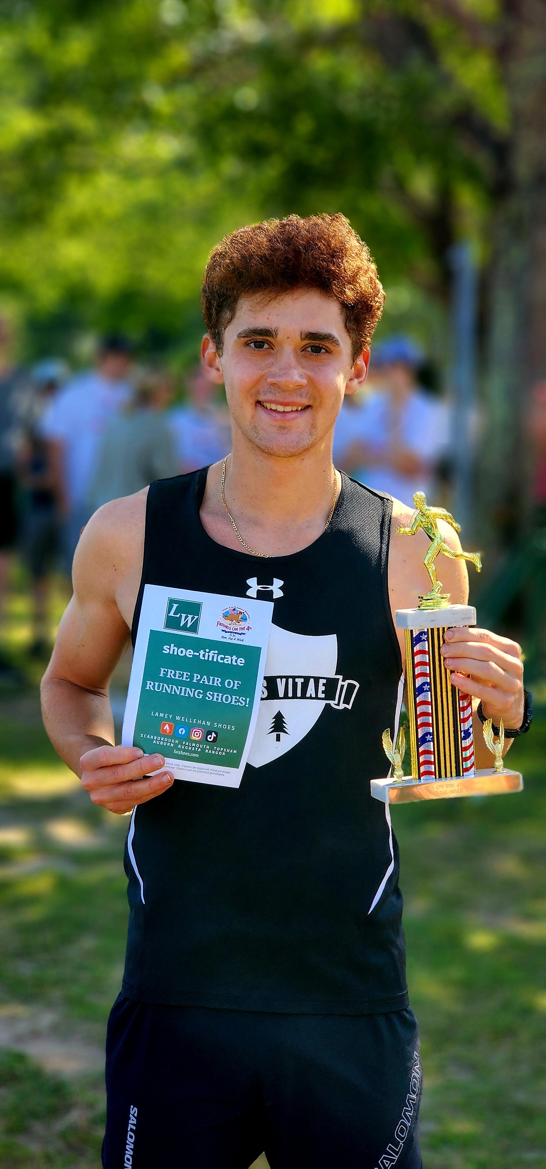 A man wearing a black tank top and shorts, holding a piece of paper that says "shoe-tificate, free pair of running shoes" and a trophy that has a figure of a runner on top, smiling at the camera