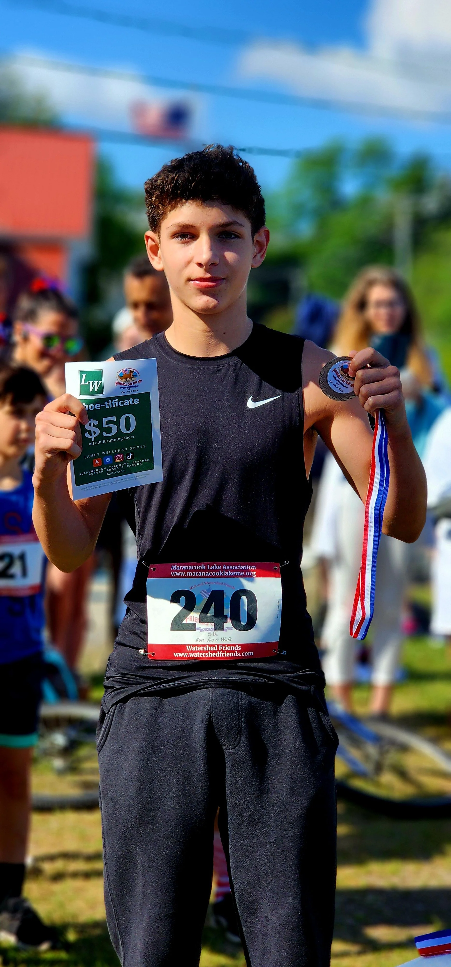 A boy wearing a black tank top and shorts, holding a certificate to Lamey Wellehan shoes and a prize medal