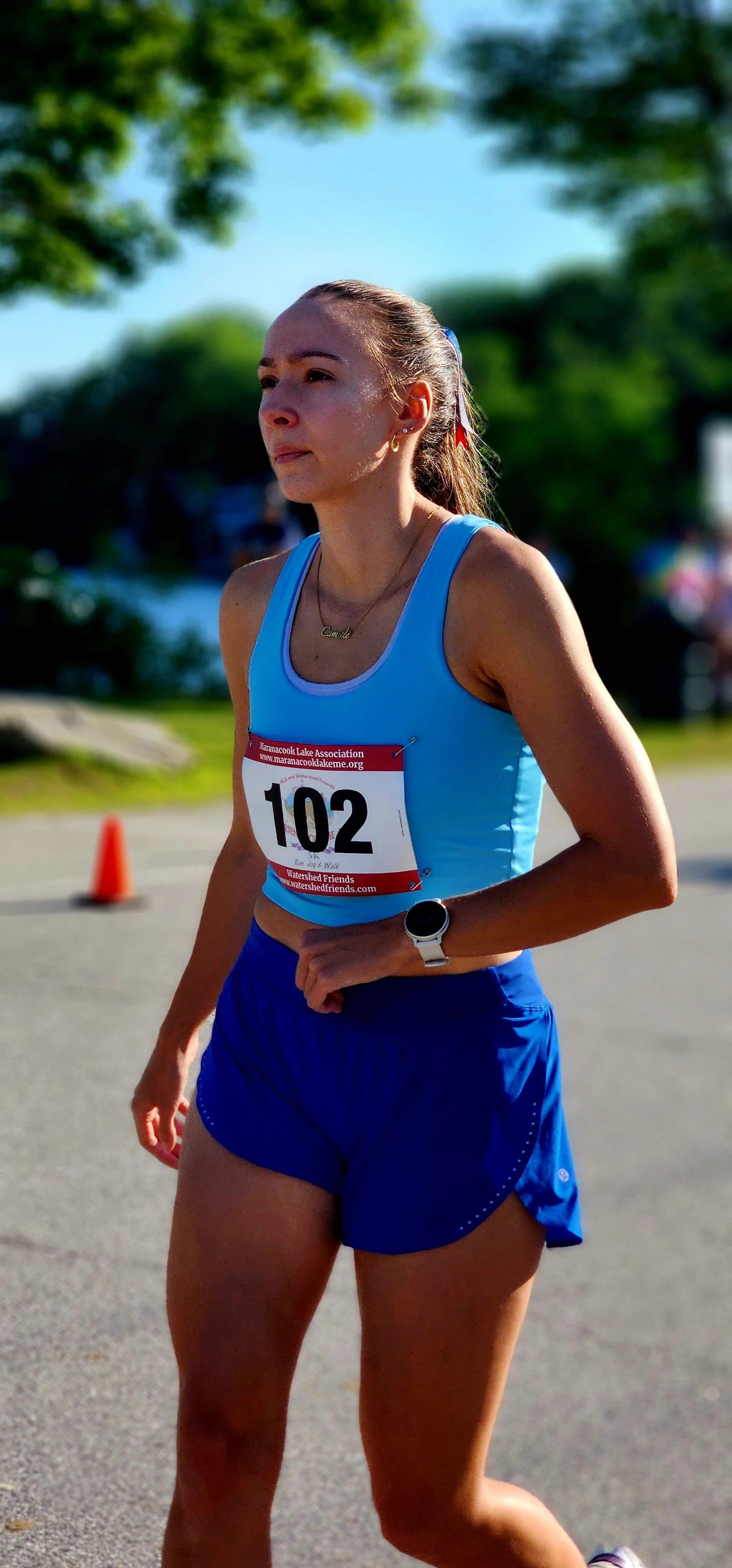 A woman, wearing a light blue tank top and blue shorts, walking with a lake in the background