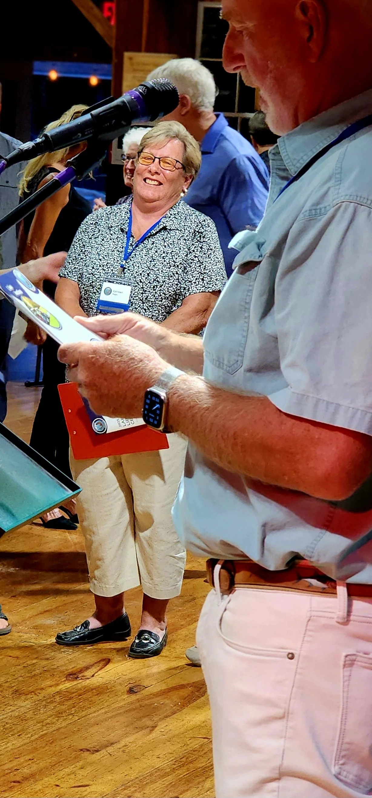 A man, wearing a light sheet short sleeve shirt and white pants, speaking into a microphone with a woman in the background laughing
