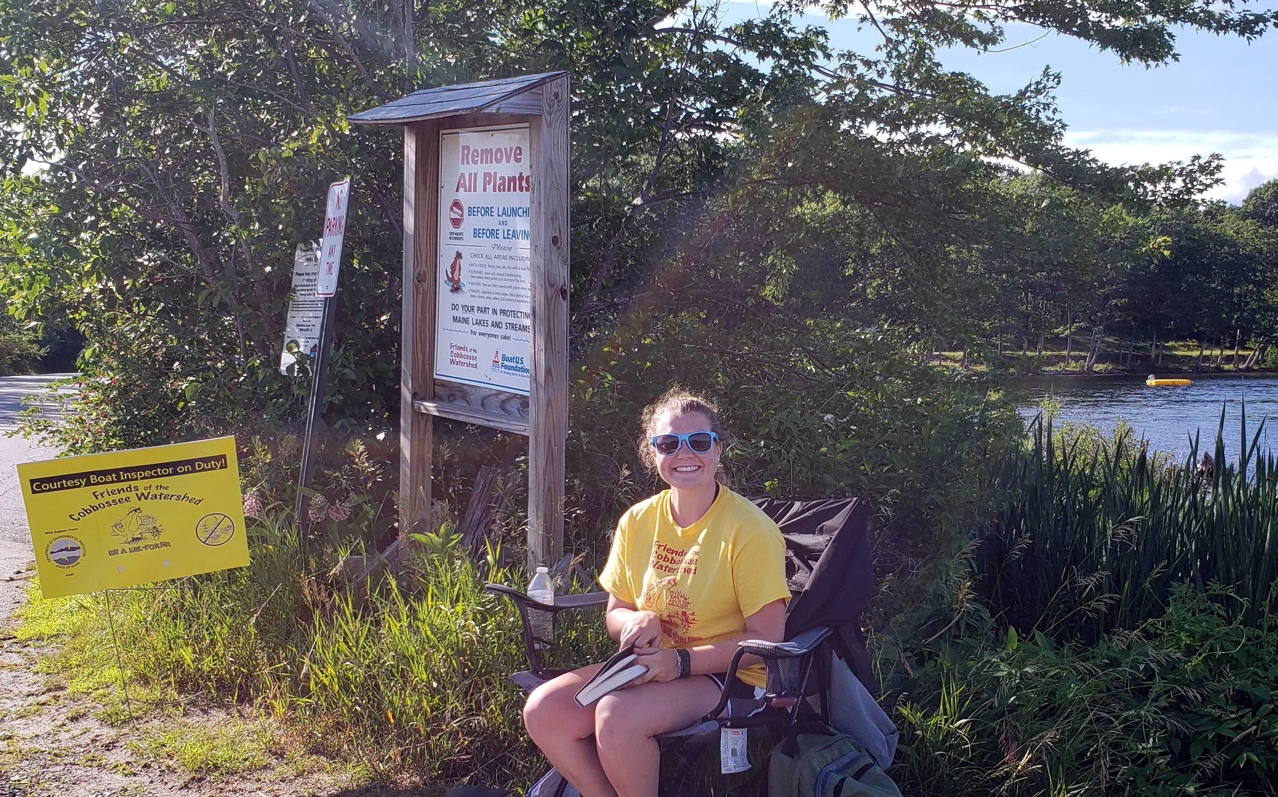 A Friends CBI in yellow staff shirt smiling at the camera and sitting in a chair next to a sign that says "Remove all plants"