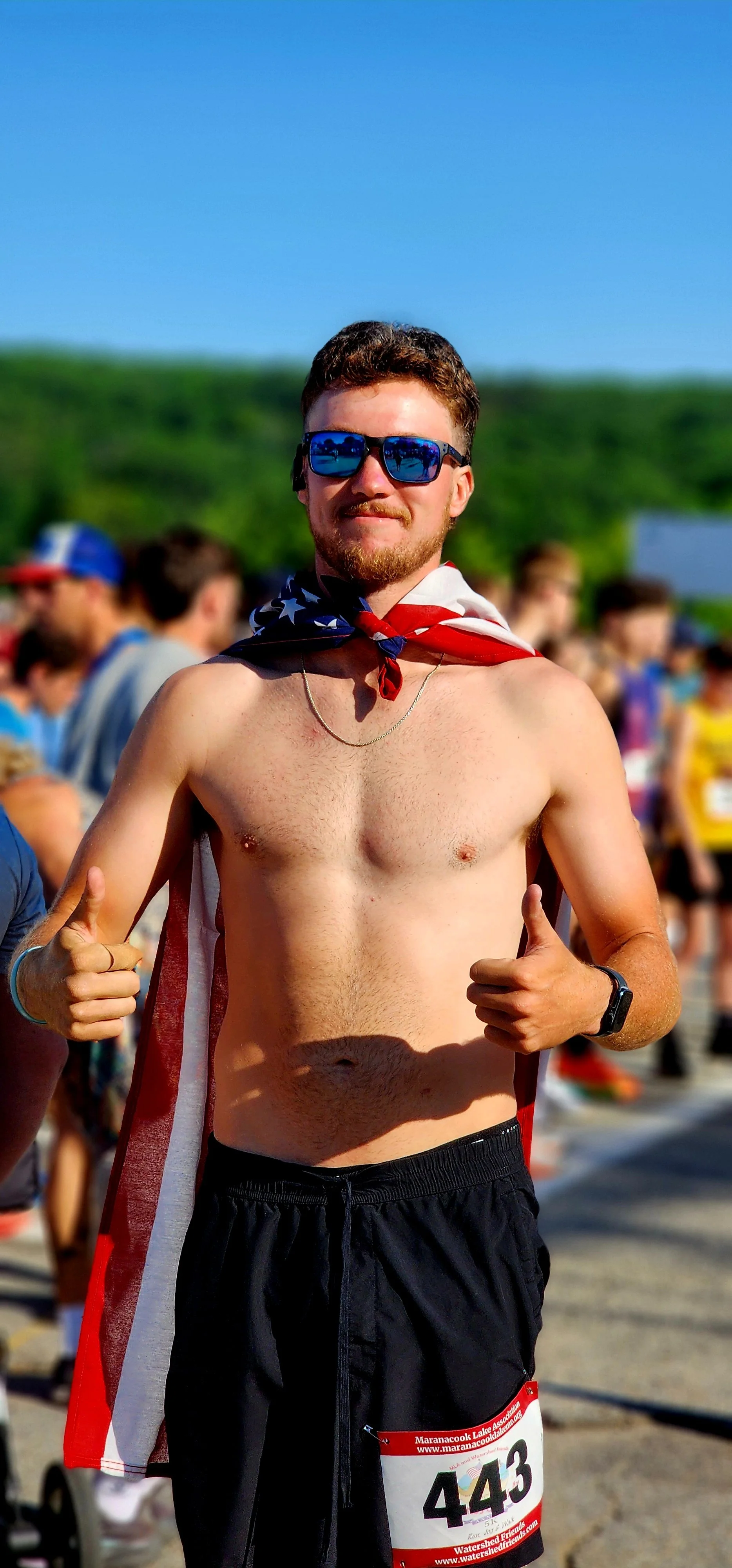 A shirtless man wearing an American flag as a cape and black shorts smiling and giving the camera two thumbs up