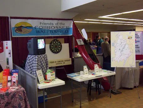 An information booth, with a "Friends of the Cobbossee Watershed" banner hanging in the wall, a map of the watershed, a table with brochures, and a red kayak leaning against the wall