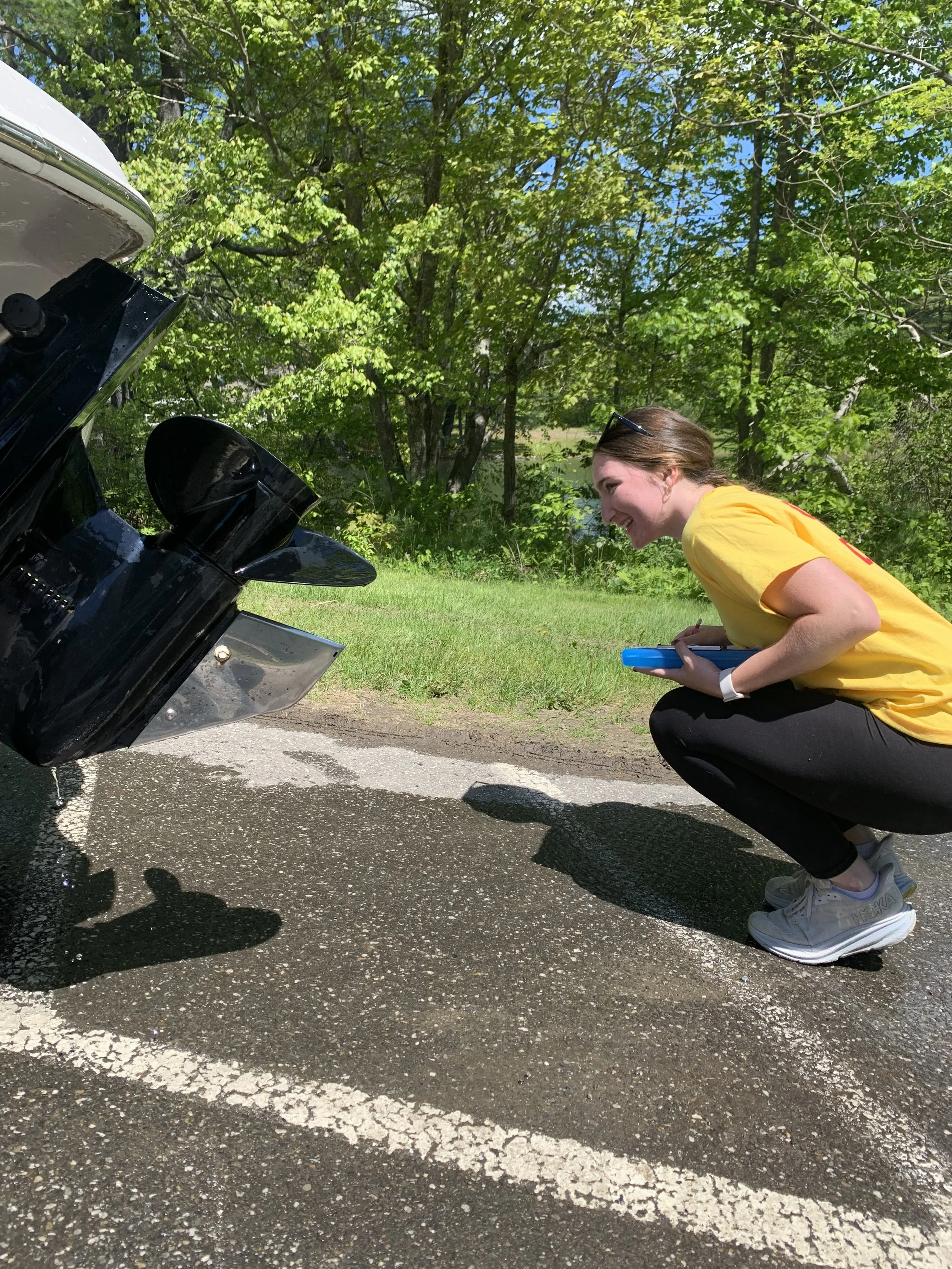 A girl wearing a yellow CBI shirt holding a clipboard and crouched down to look at the prop of a boat on a trailer