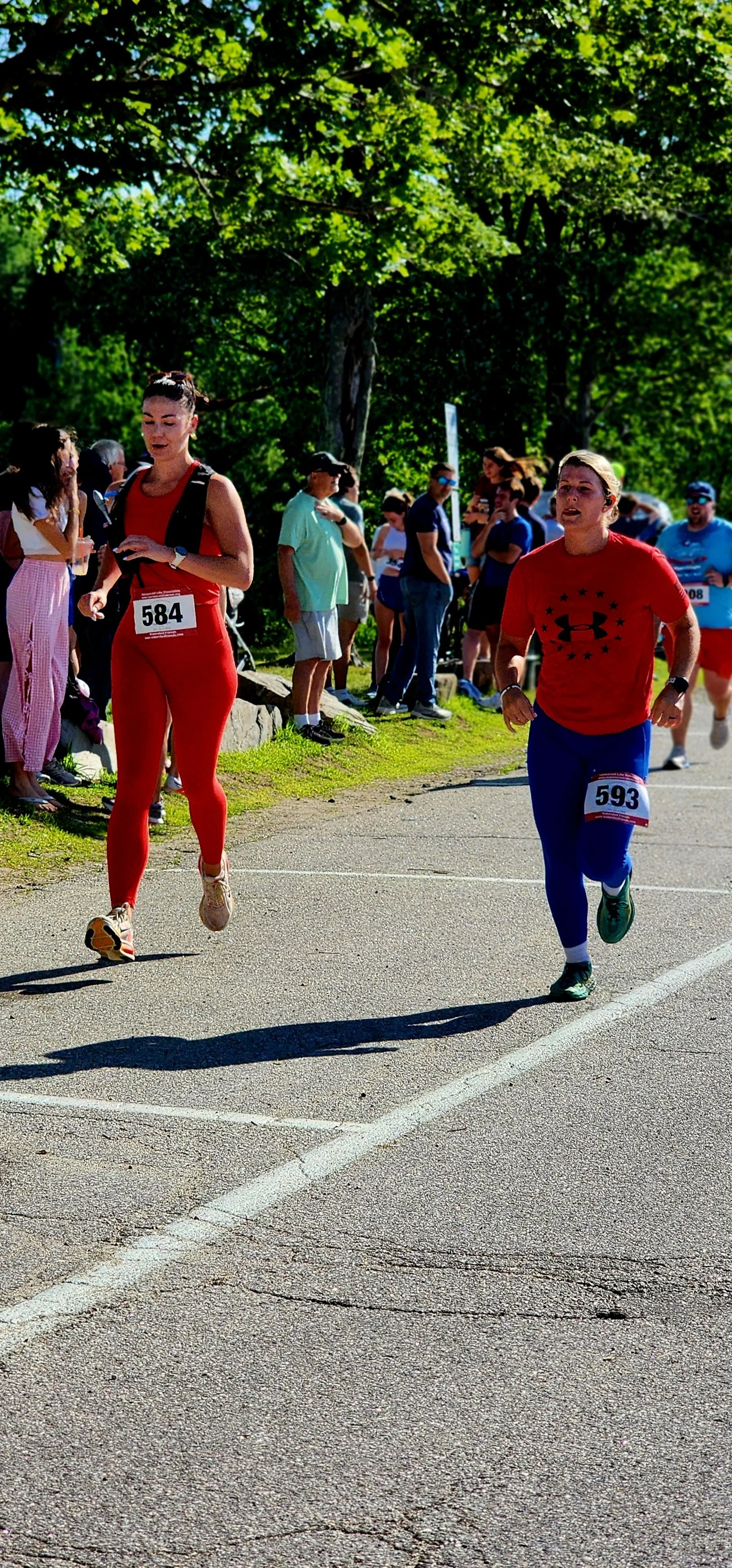 Two women running, one wearing a red tank top and red leggings the other wearing a red t-shirt and blue shorts, with a crowd standing in the background
