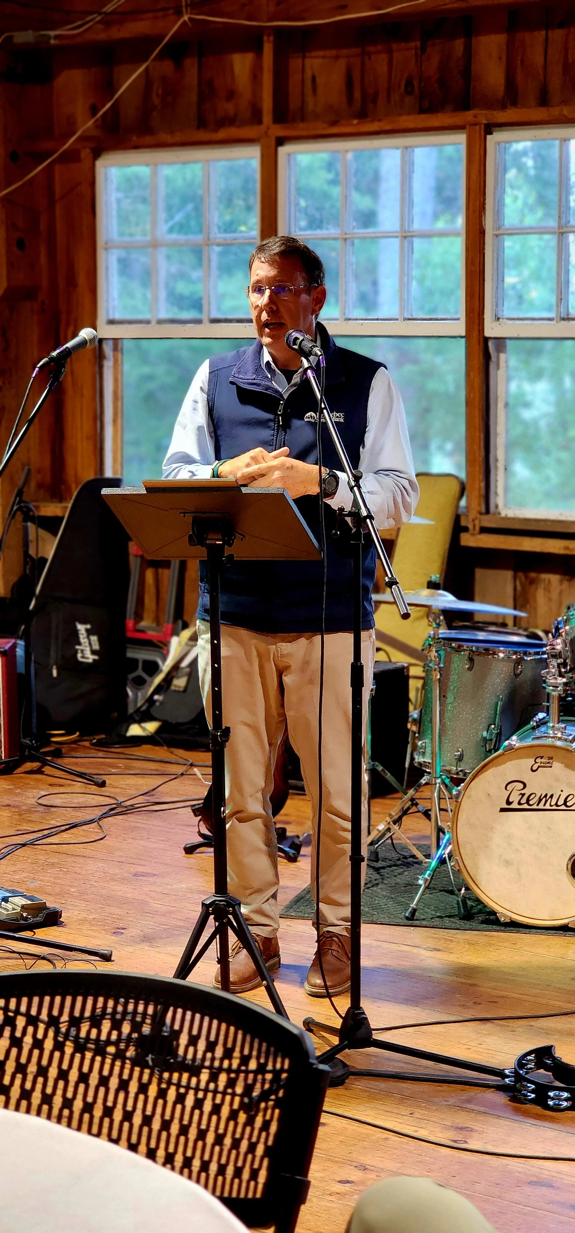 A man, wearing a dark blue vest over light blue long sleeve with khakis, speaking into a microphone with a stand in front of him