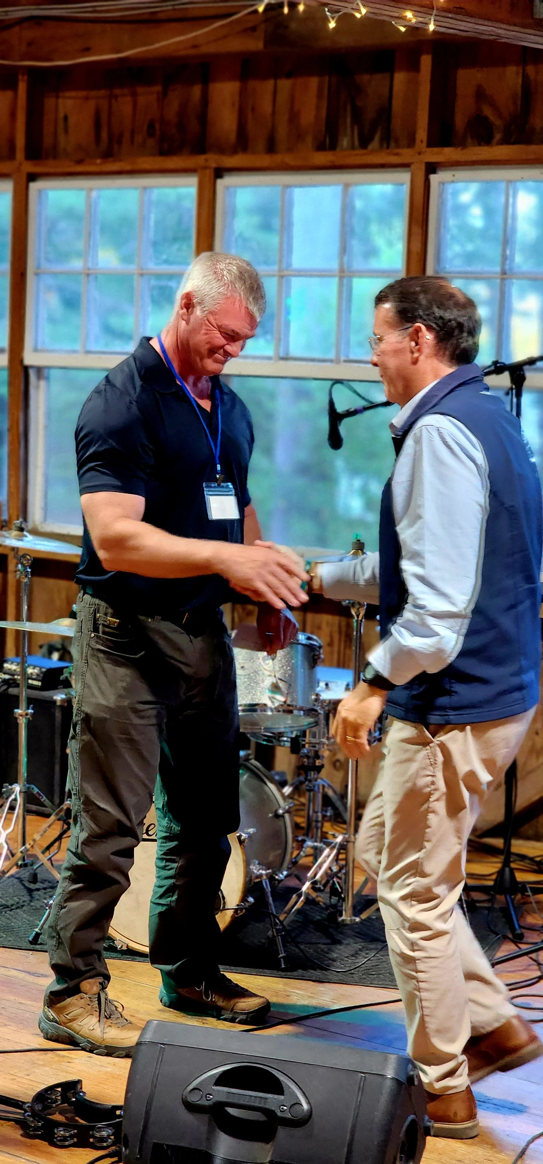 Two men, one wearing a black polo and dark grey pants and the other wearing a dark blue vest over a blue long sleeve and khakis, speaking by a microphone