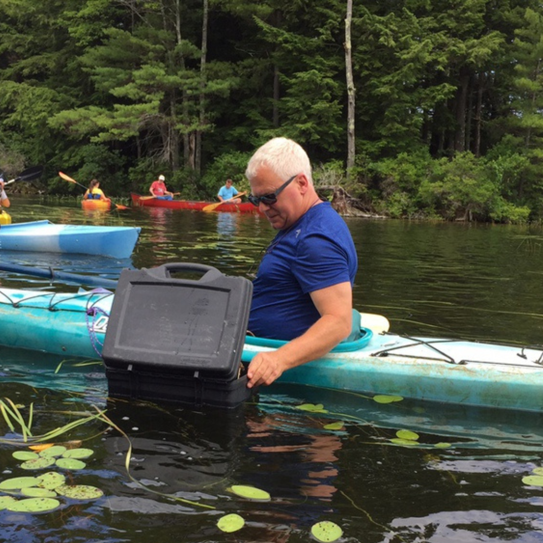 A man in a kayak wearing a blue shirt and sunglasses looking down into a trunk style viewing scope