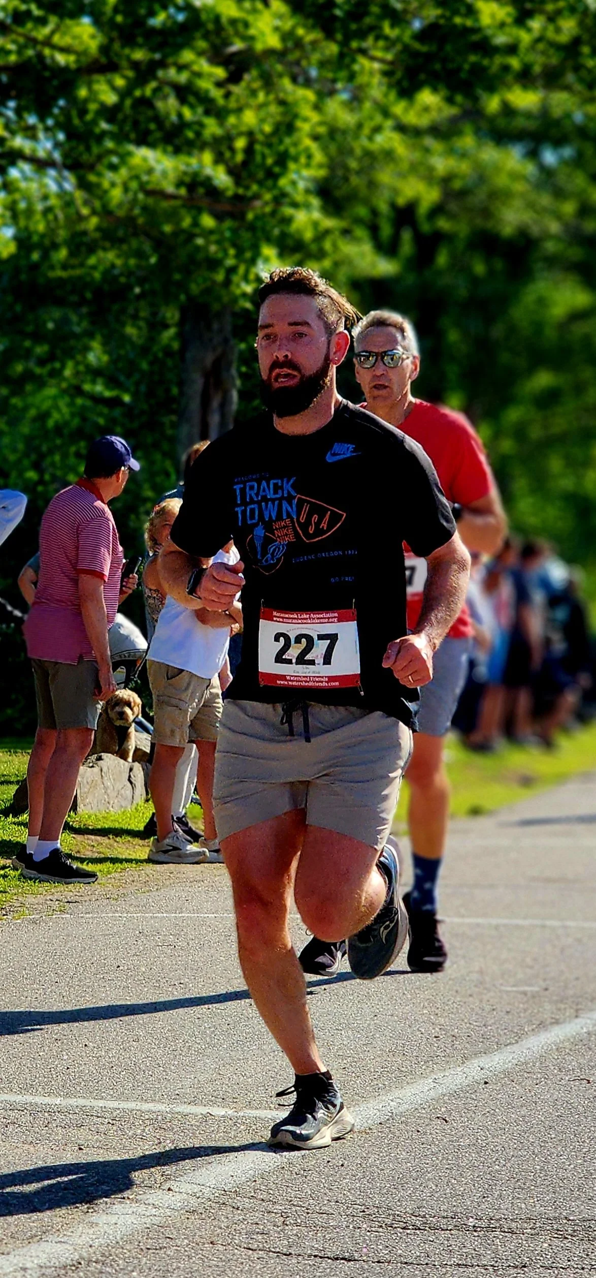A man wearing a black t-shirt and grey shorts running with a crowd standing in the background