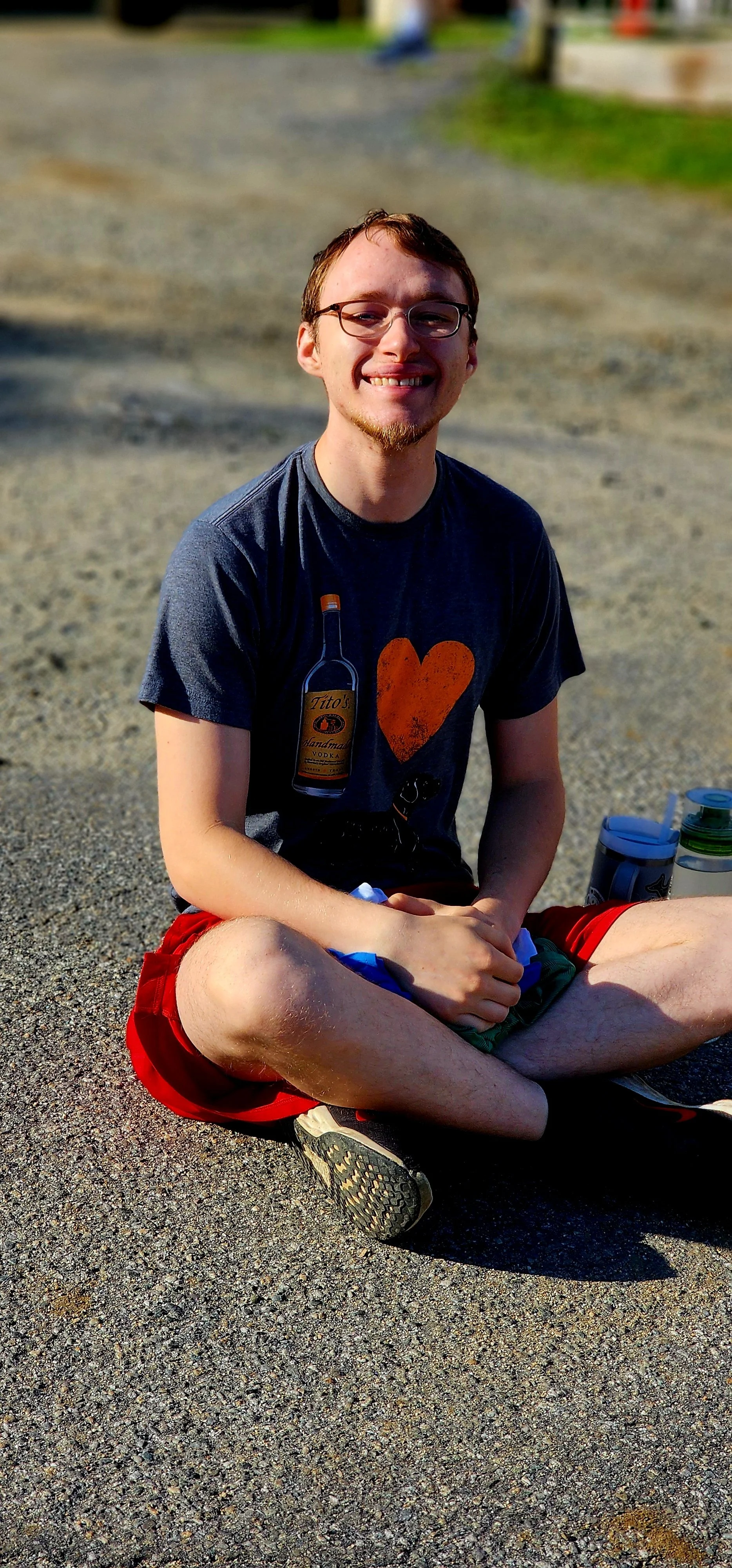 A man, wearing a dark grey shirt and red shorts, sitting cross legged on the ground, smiling at the camera