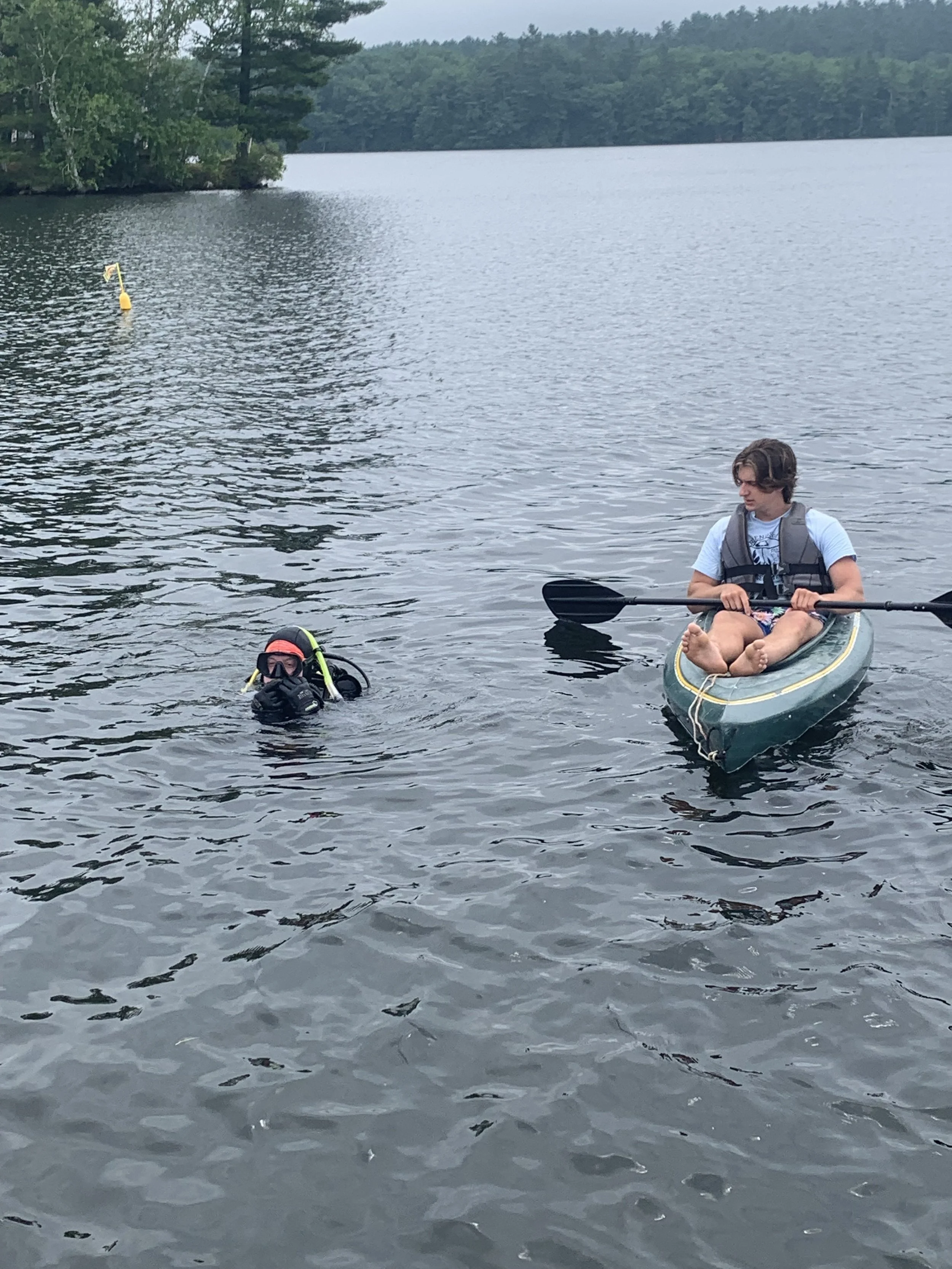 A diver with their head out of water and hands adjusting their gear, with a man in a green kayak wearing a blue t-shirt watching the diver