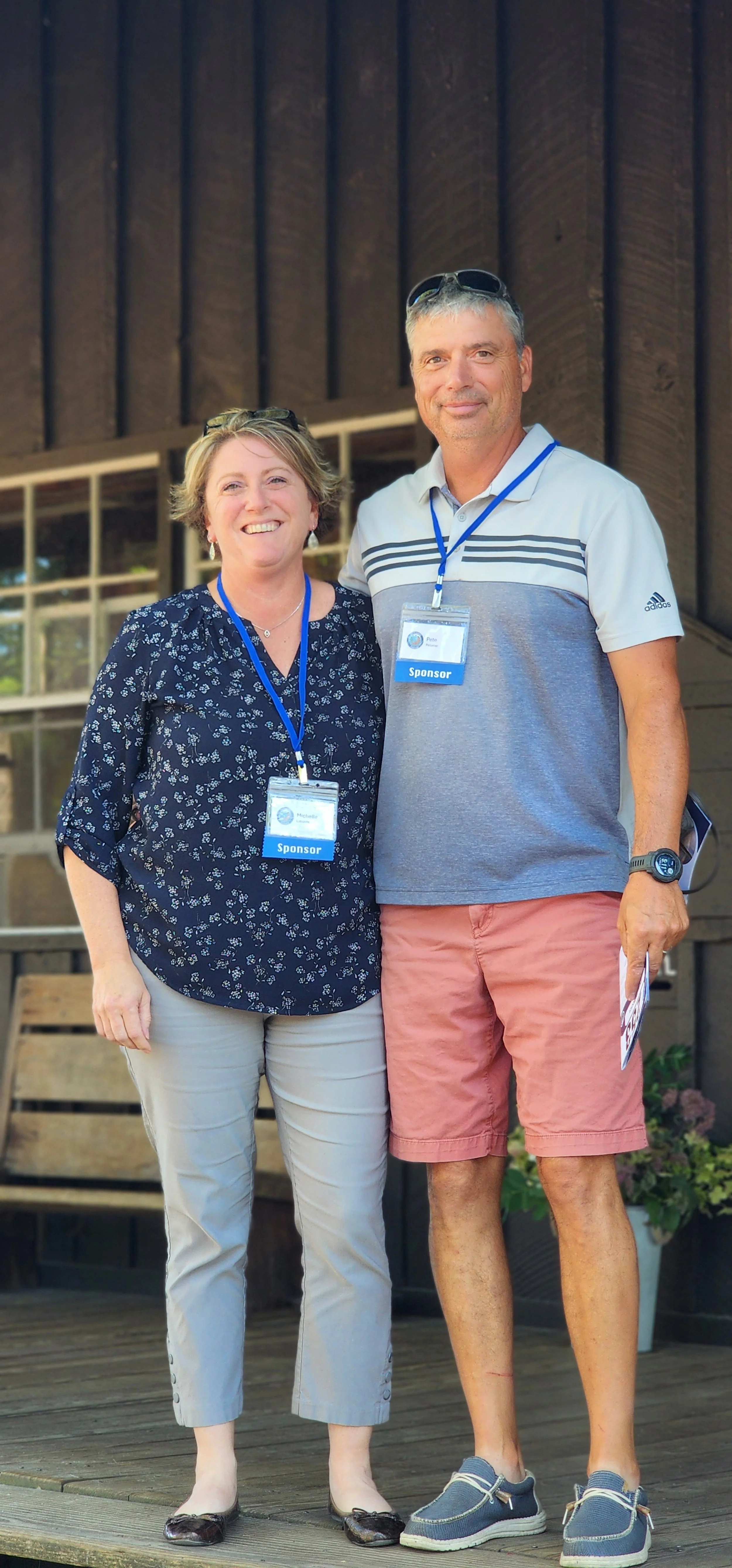 A woman, wearing a dark blue shirt and grey pants, and a man, wearing a grey striped polo shirt and salmon shorts, standing on a porch