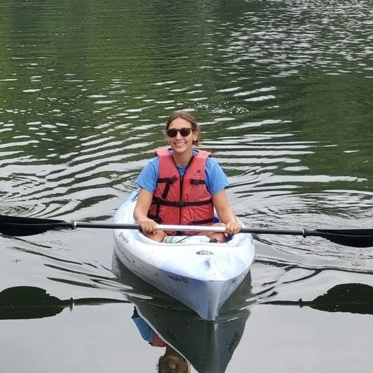 A person in a kayak wearing a blue shirt, red life jacket, and sunglasses smiling at the camera
