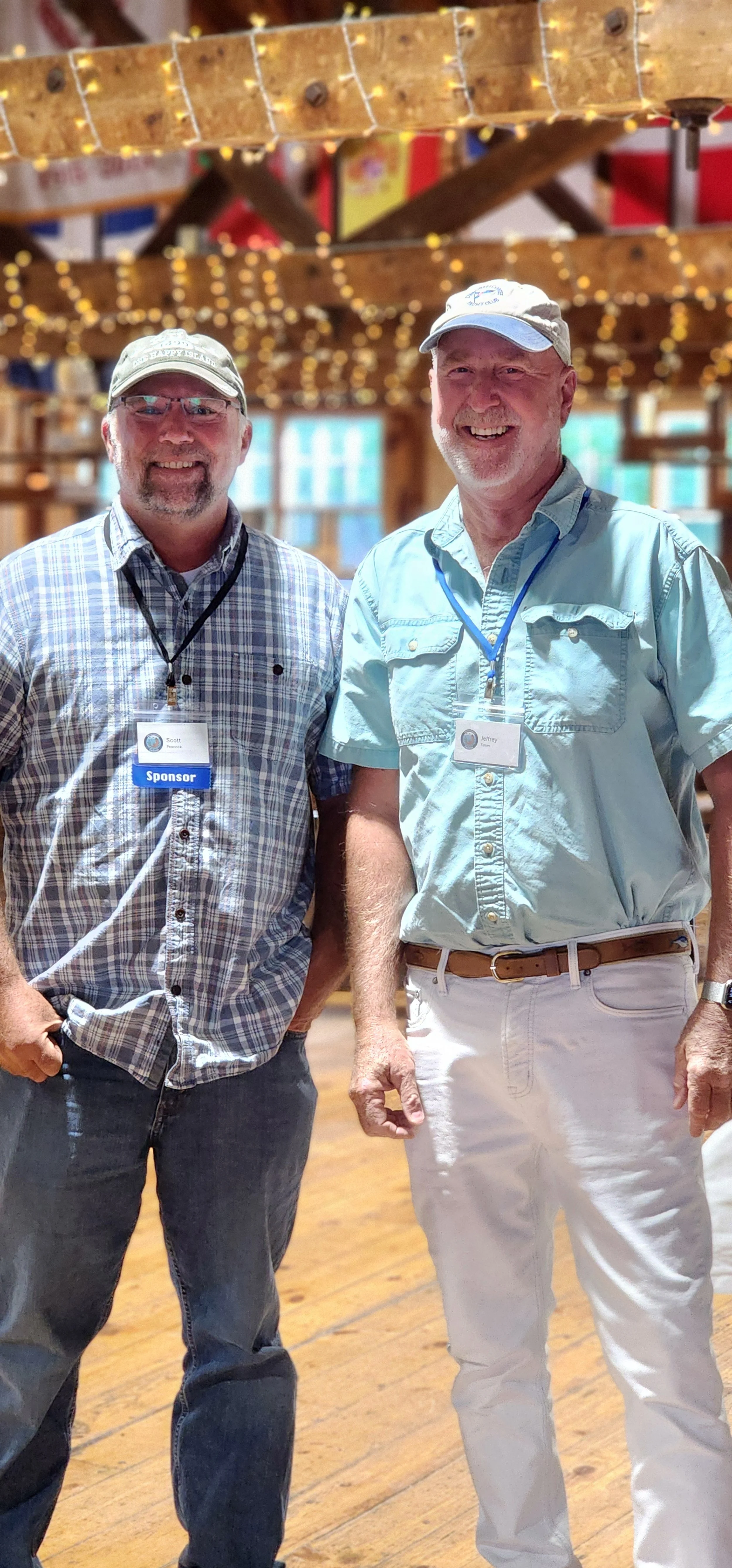 Two men, one wearing a blue and white plaid shirt and jeans, the other wearing a green shirt and white pants, smiling at the camera