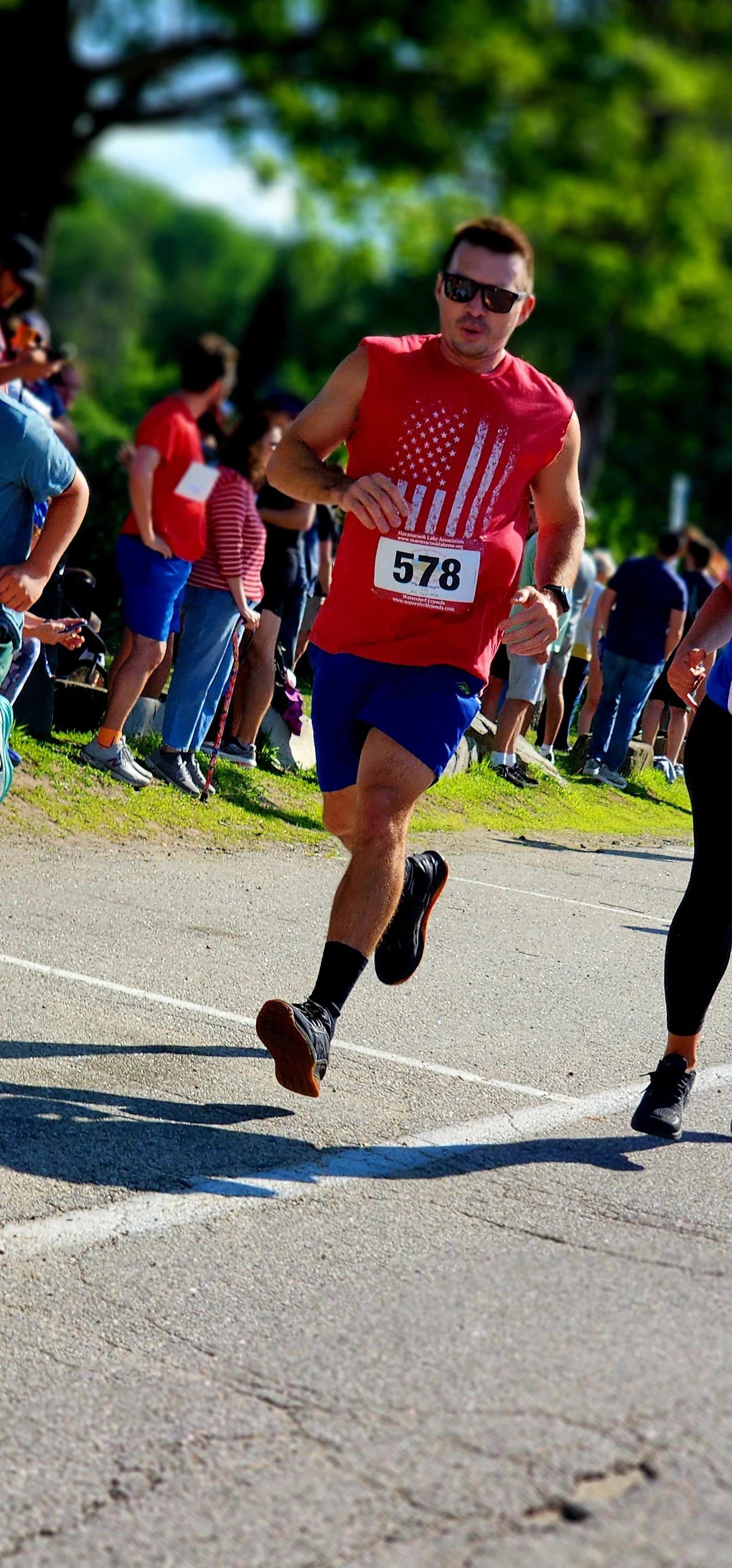 A man wearing a red tank top and blue shorts running with a crowd standing in the background