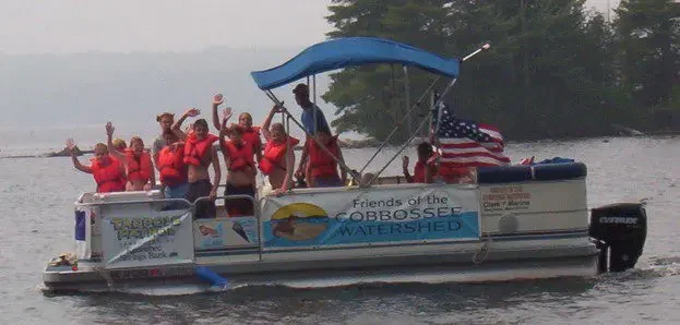 A pontoon boat with a "Friends of the Cobbossee Watershe" banner and group of people waving at the camera