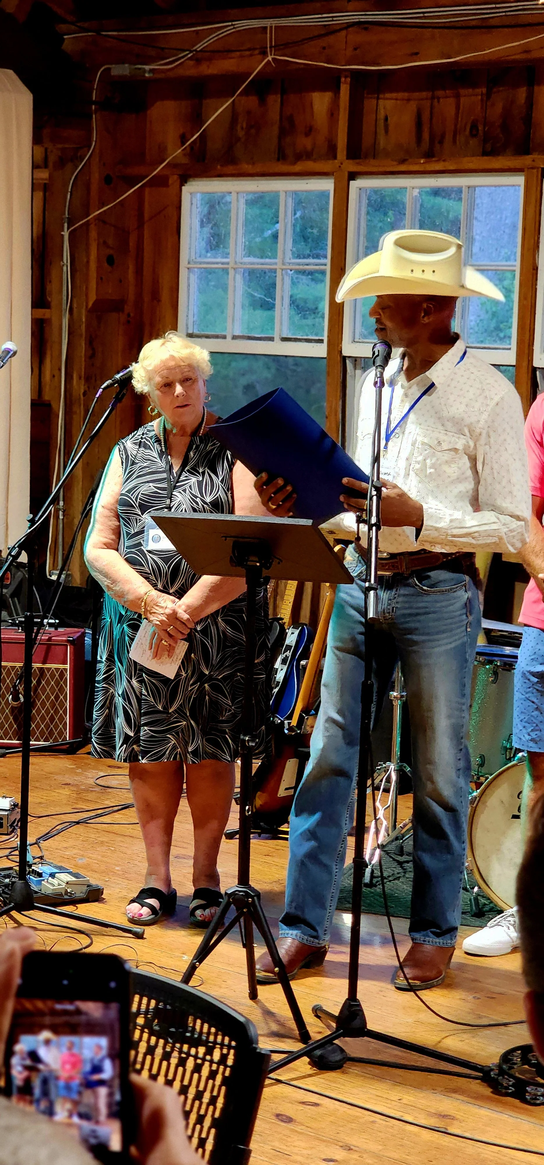 A woman, wearing a black dress with white patterns, and a man, wearing a white cowboy hat with a white shirt and jeans, the man is speaking into a microphone