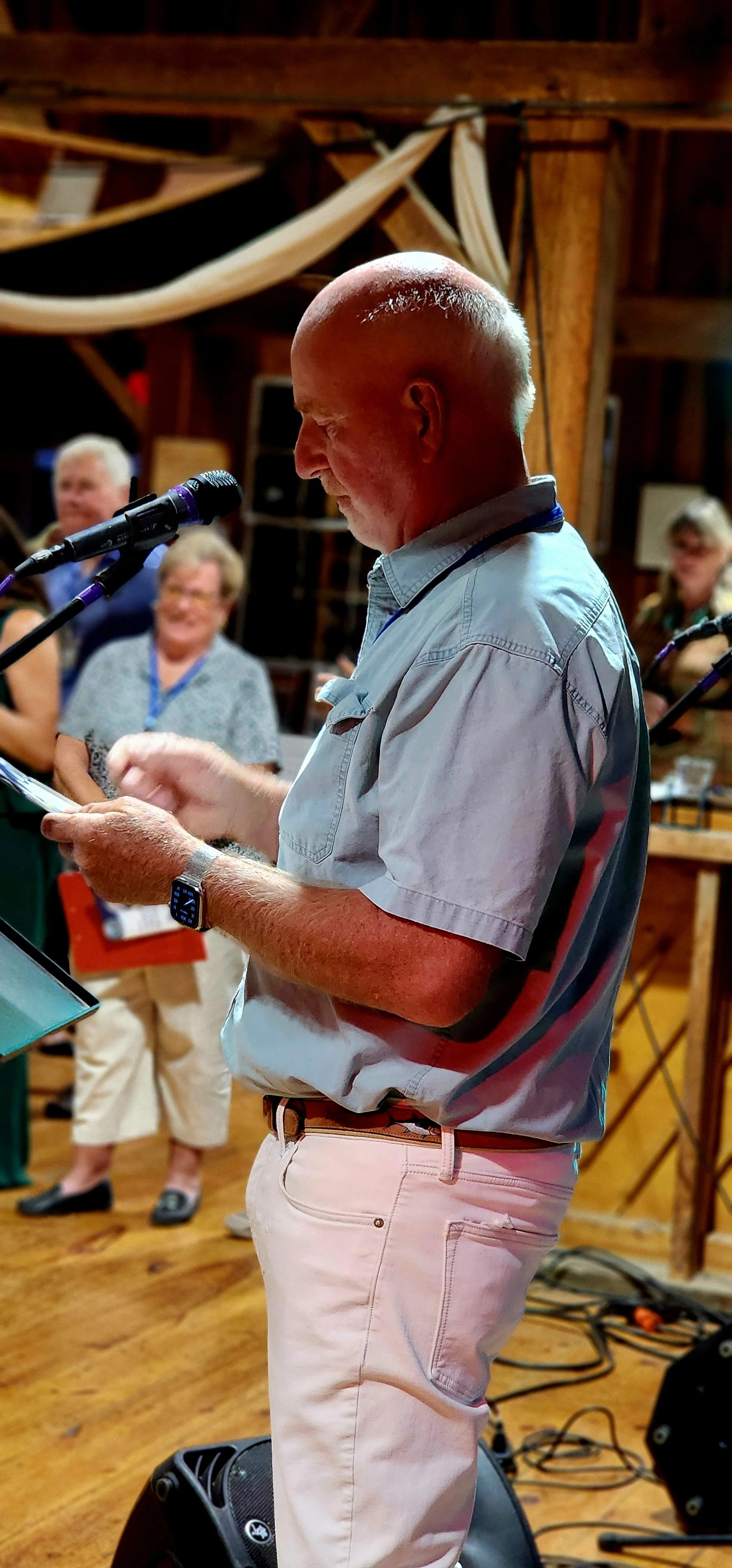 A man, wearing a light green short sleeve and white pants, speaking into a microphone