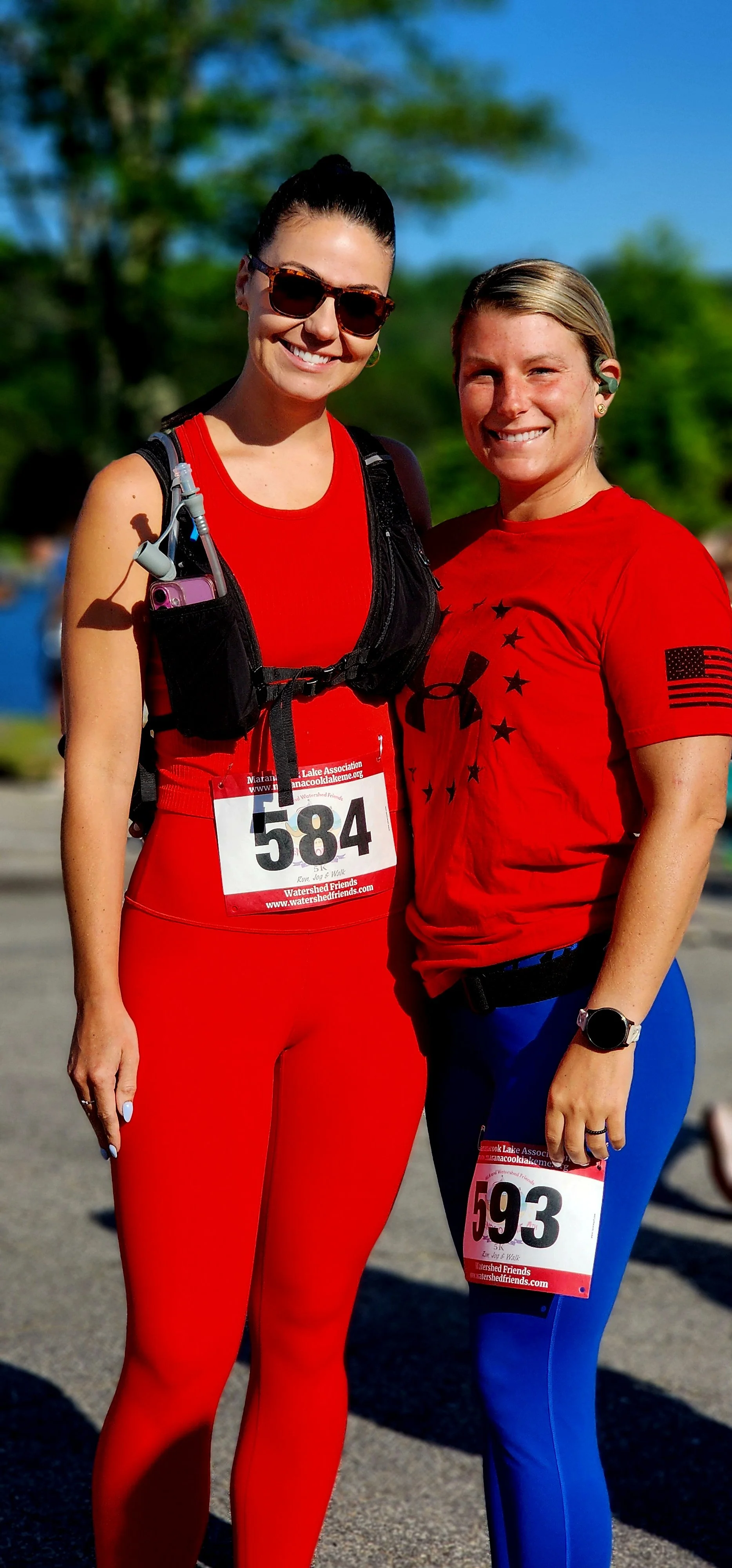 Two women, one wearing a red tank top and red leggings and the other wearing a red t-shirt and blue leggings, smiling at the camera