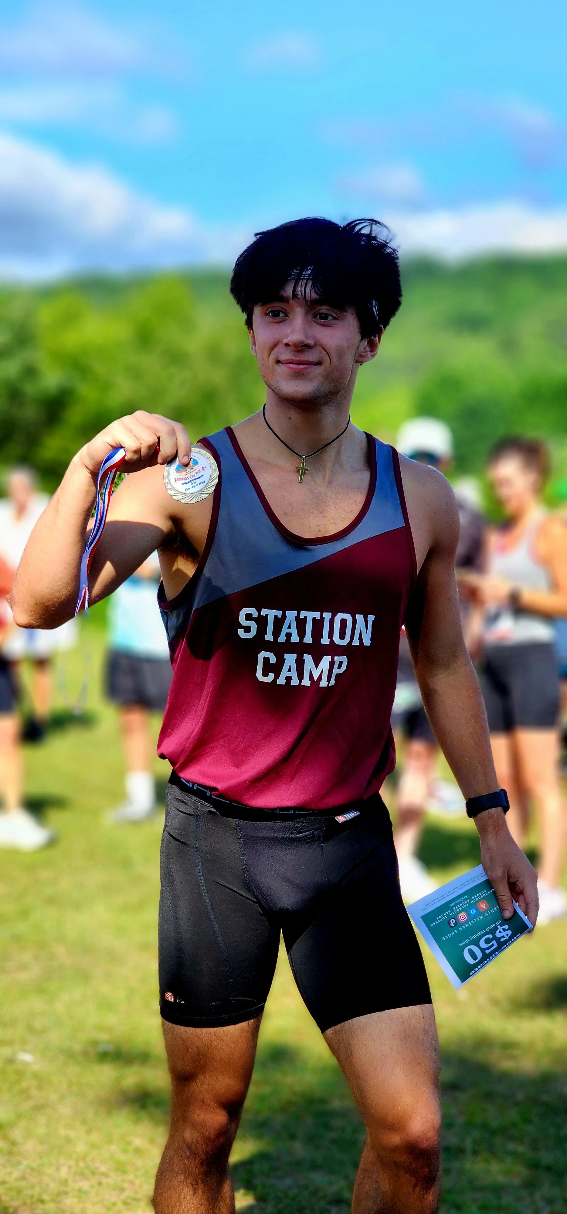 A boy, wearing a maroon and grey tank top and black shorts holding up an award medal