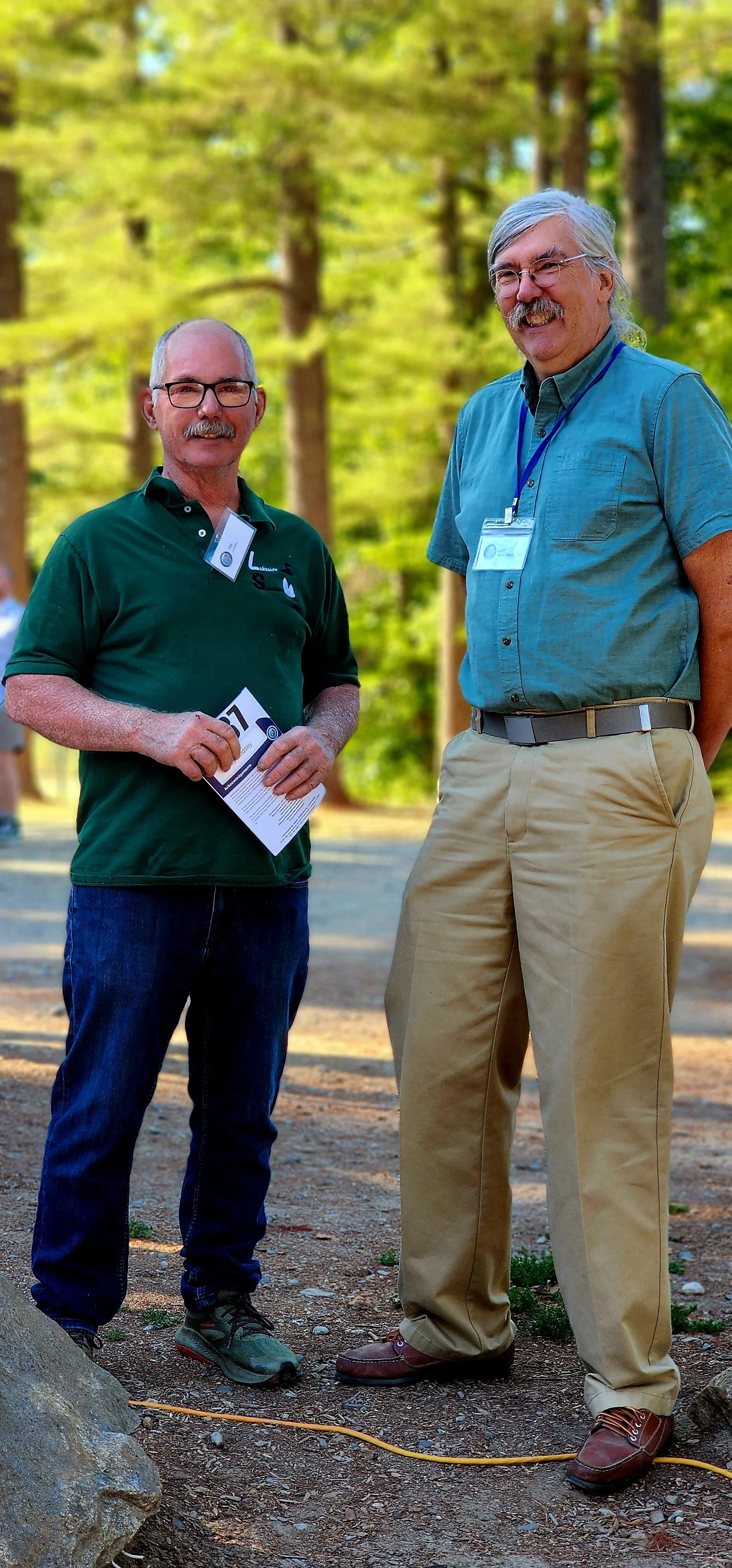 Two men, one wearing a green polo and jeans the other wearing a blue green short sleeve button up shirt and khakis, smiling at the camera