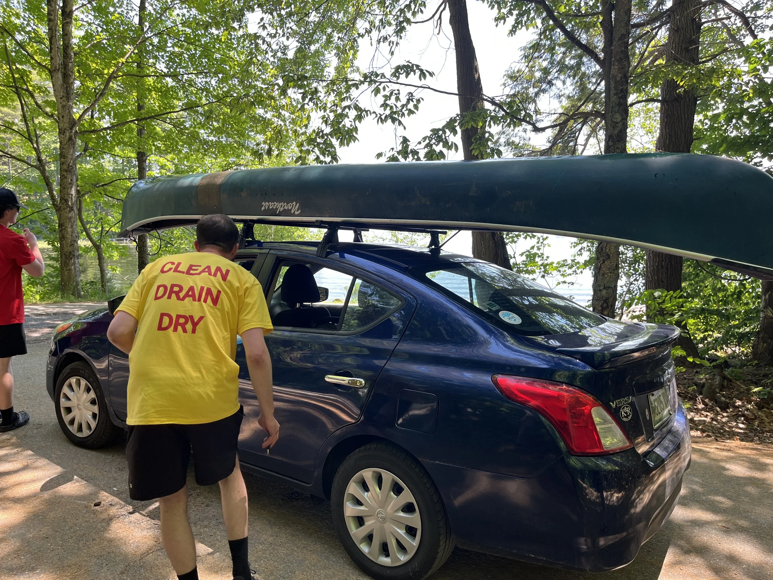 A man facing away from the camera, wearing a yellow shirt that says "Clean Drain Dry" on the back, leaning down to speak in the window of a blue sedan that has a green canoe on its roof