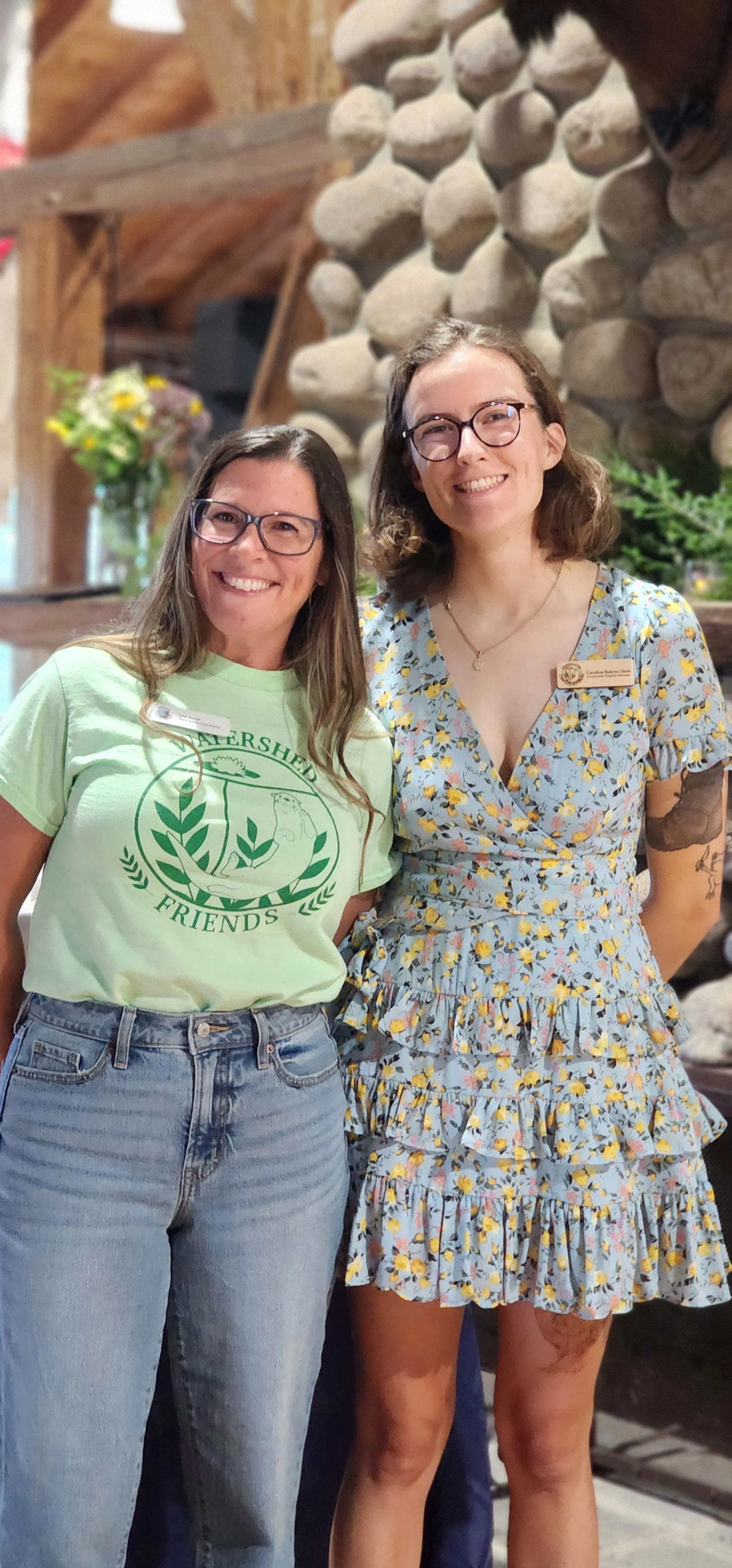 Two people, the one on the left wearing a green t-shirt and jeans, the one on the right wearing a light blue and yellow floral patterned dress, smiling at the camera
