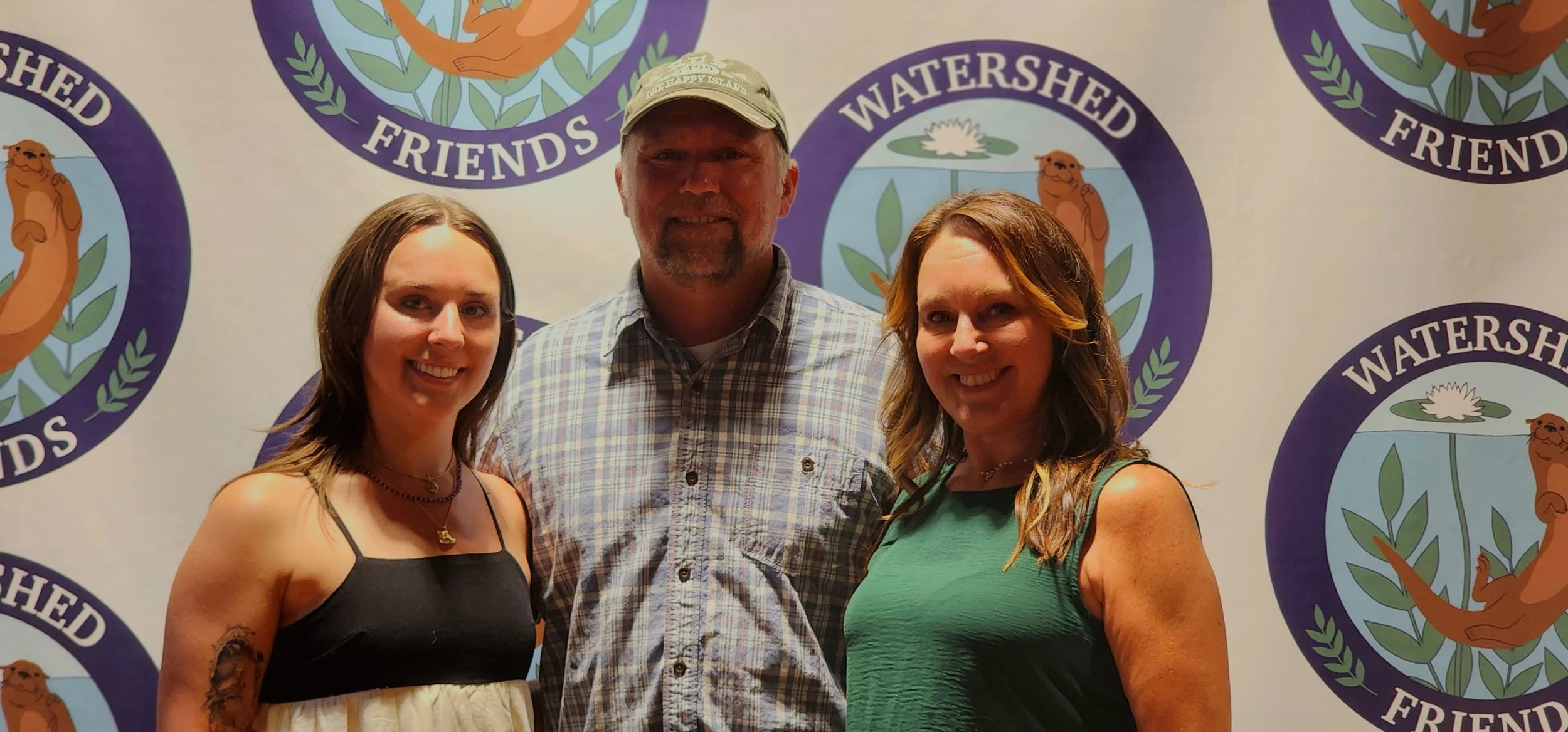 Two women, one wearing a black and white dress the other wearing a green dress, with a man, wearing a tan ball cap and blue plaid shirt, smiling at the camera