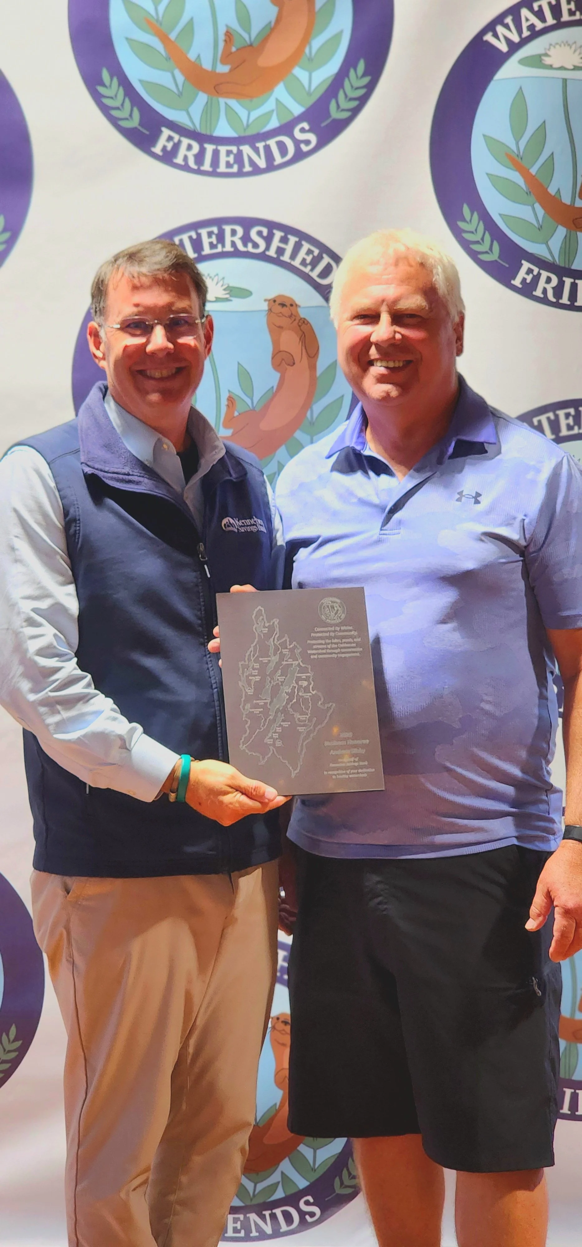 Two men, one wearing a navy vest over a blue shirt and khakis holding an award, the other wearing a blue polo and black shorts, smiling in front of a backdrop with the Watershed Friends logo
