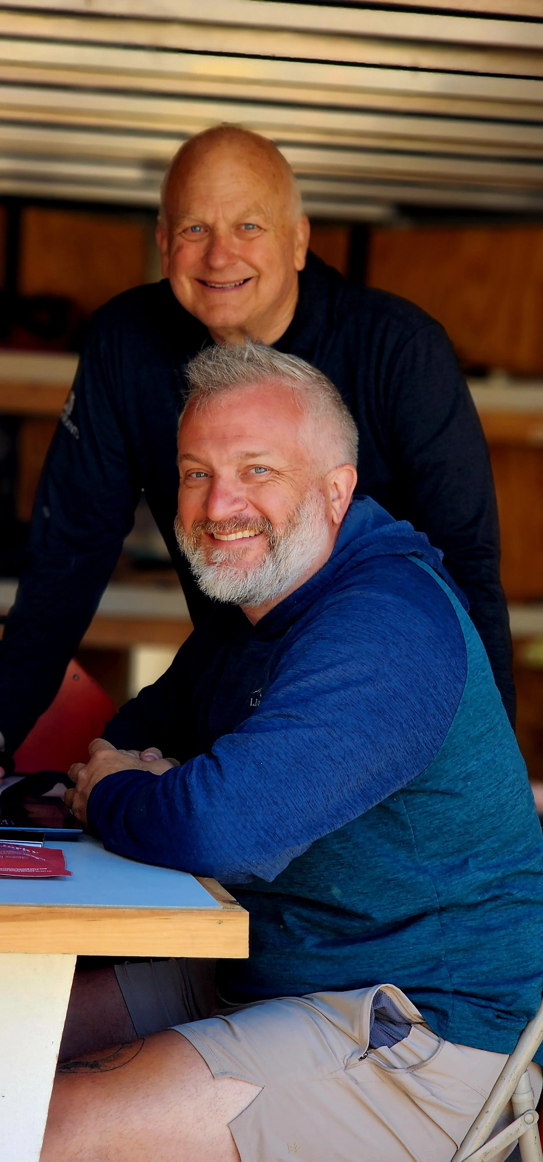 Two men, one in back wearing a black long sleeve shirt and one in front wearing a blue long sleeve shirt and khaki shorts, smiling at the camera