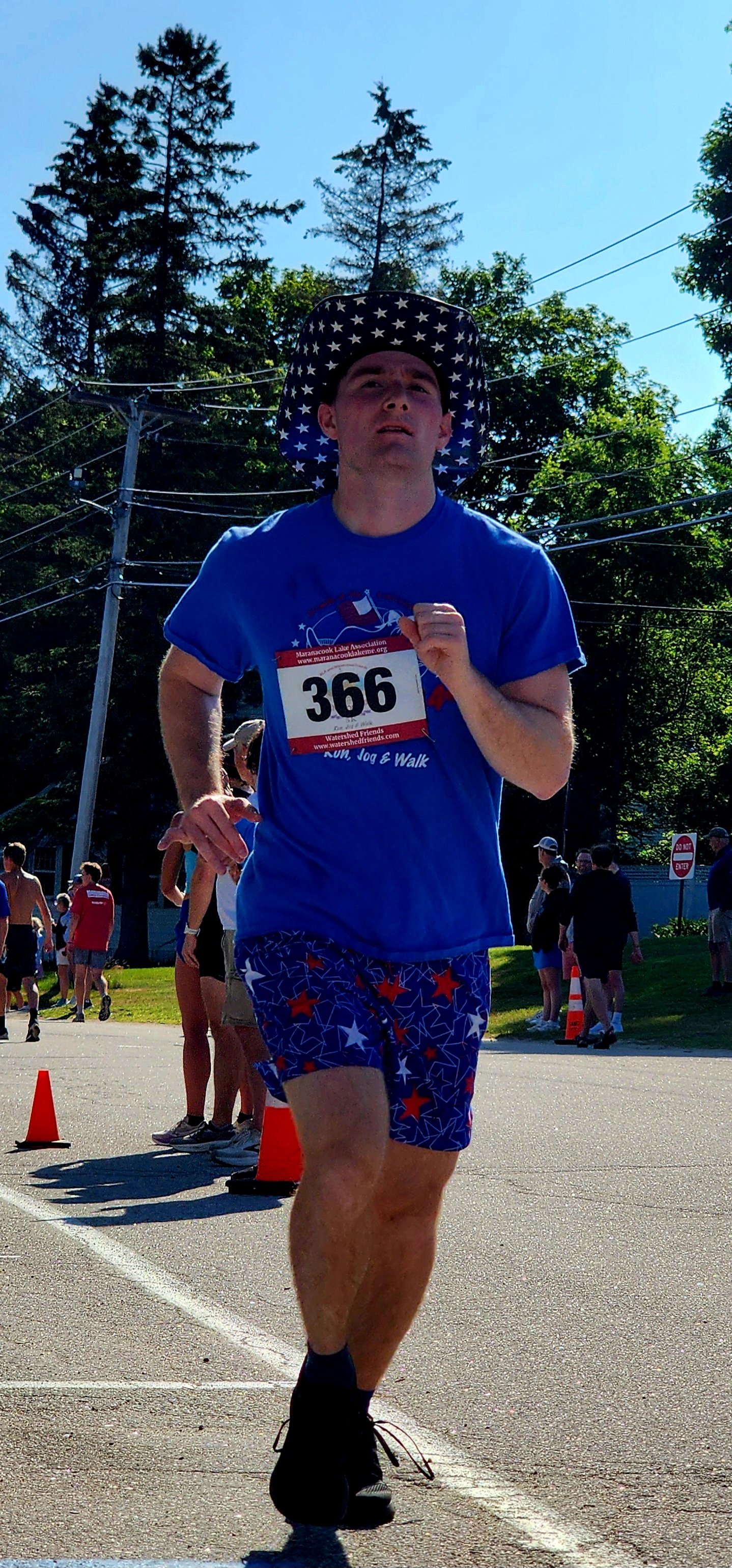 A man wearing a patriotic cowboy hat, blue t-shirt, and blue shorts running