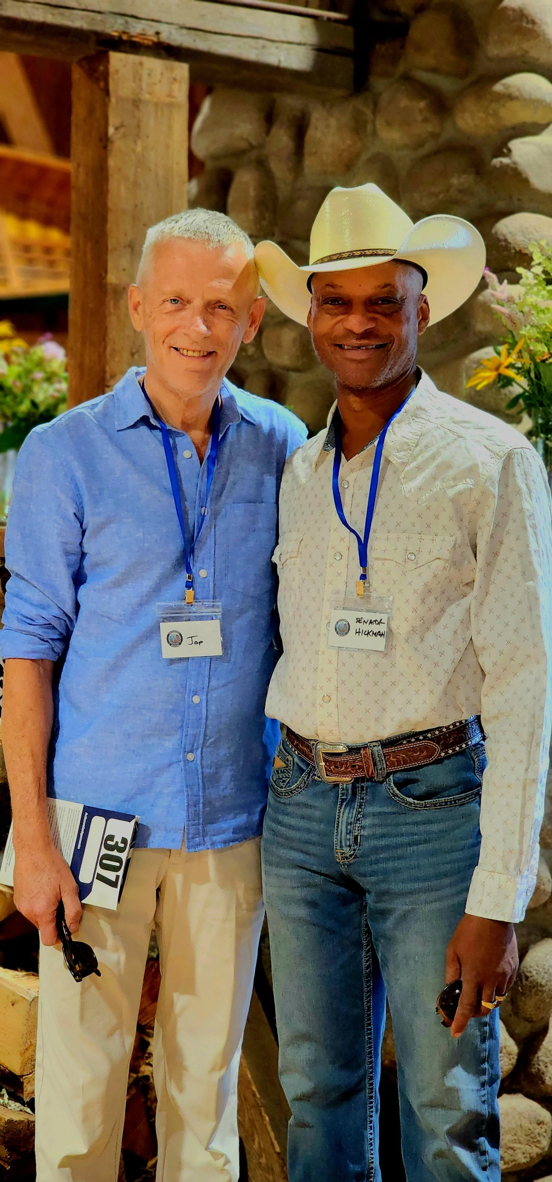 Two men smiling at the camera, the one on the left wearing a light blue button up shirt and khakis, the one on the right wearing a white cowboy hat, white button up shirt, and jeans