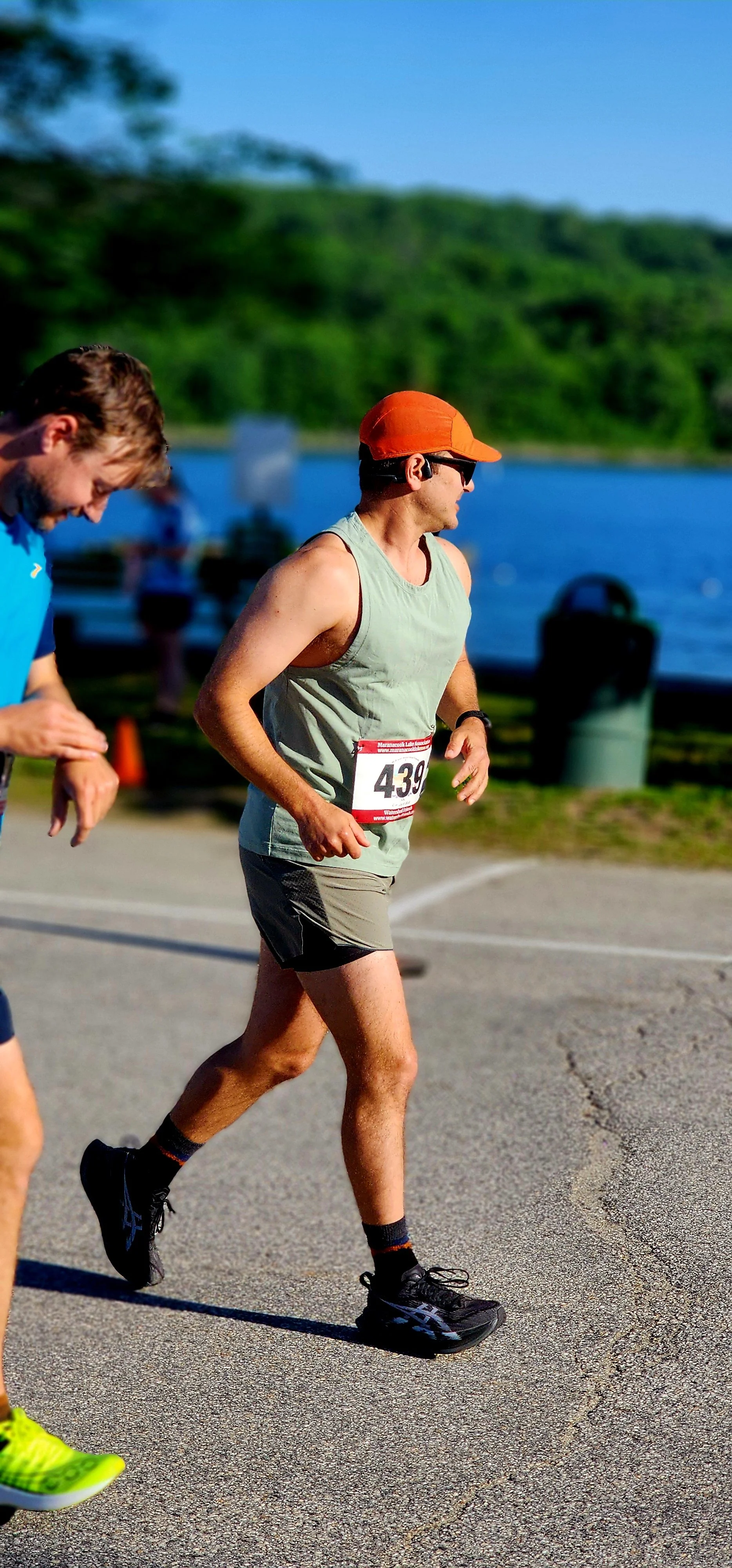 A man, wearing a light grey tank top, grey shorts and an orange ball cap, jogging with a lake in the background